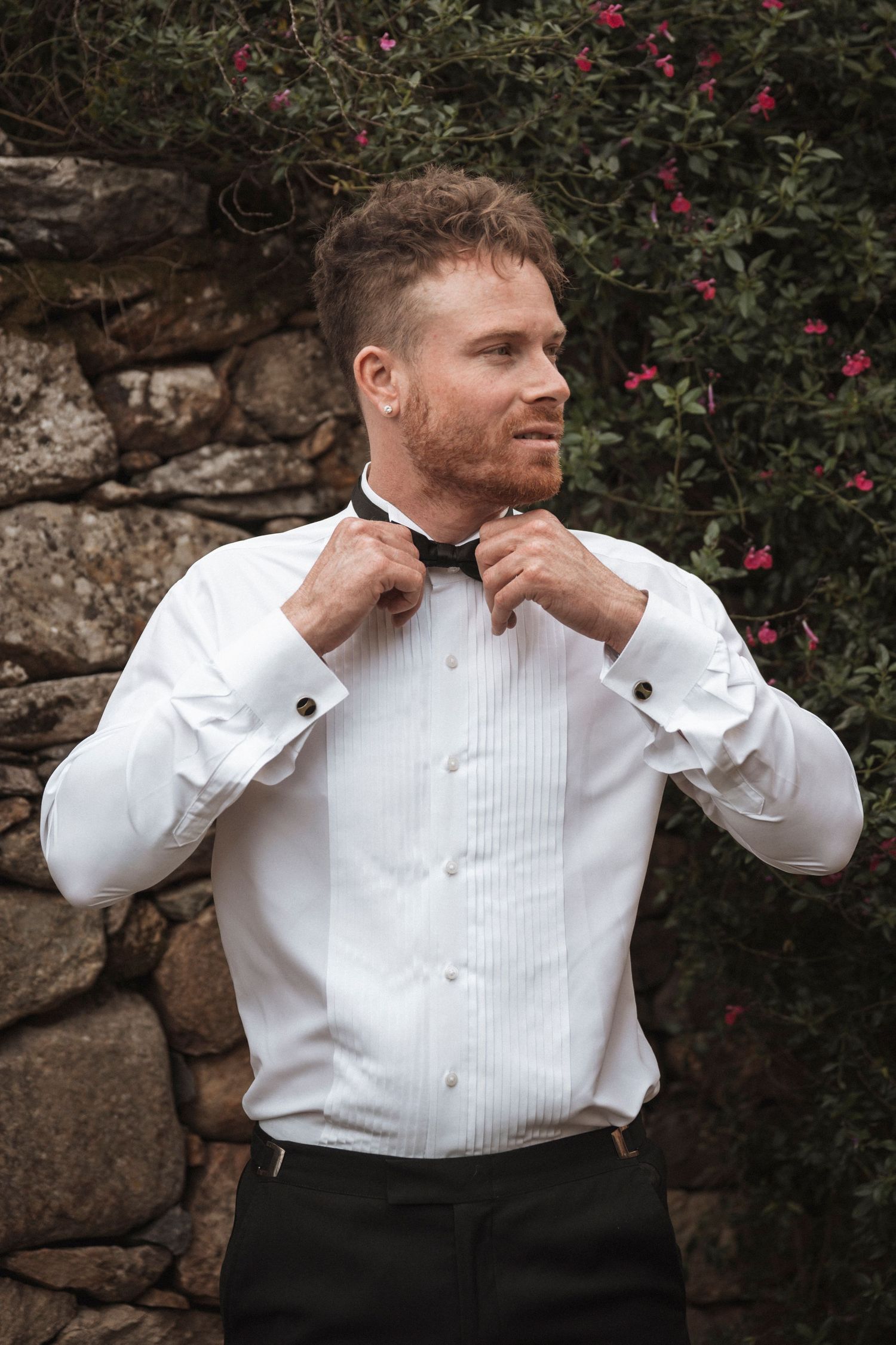Person adjusting white tuxedo shirt collar against stone wall with red flowering bushes.