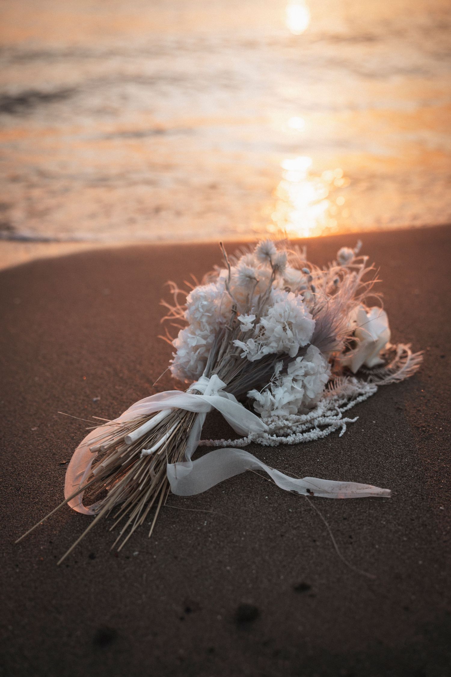 A discarded white floral bouquet lies on dark sand during golden hour at the beach.