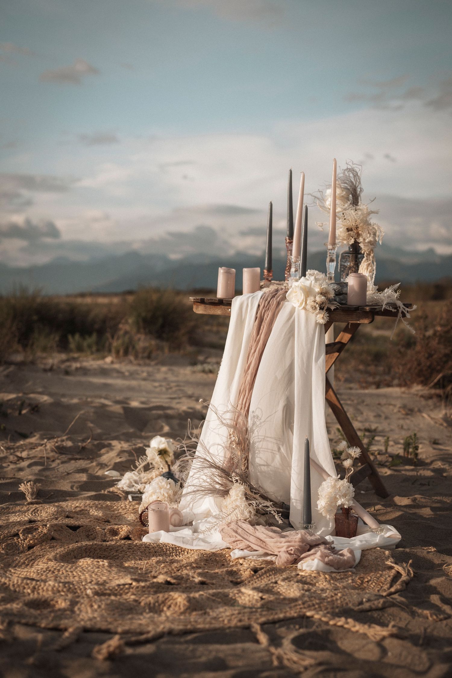 Romantic wedding decor setup with candles and fabric on desert sand with mountains in background.