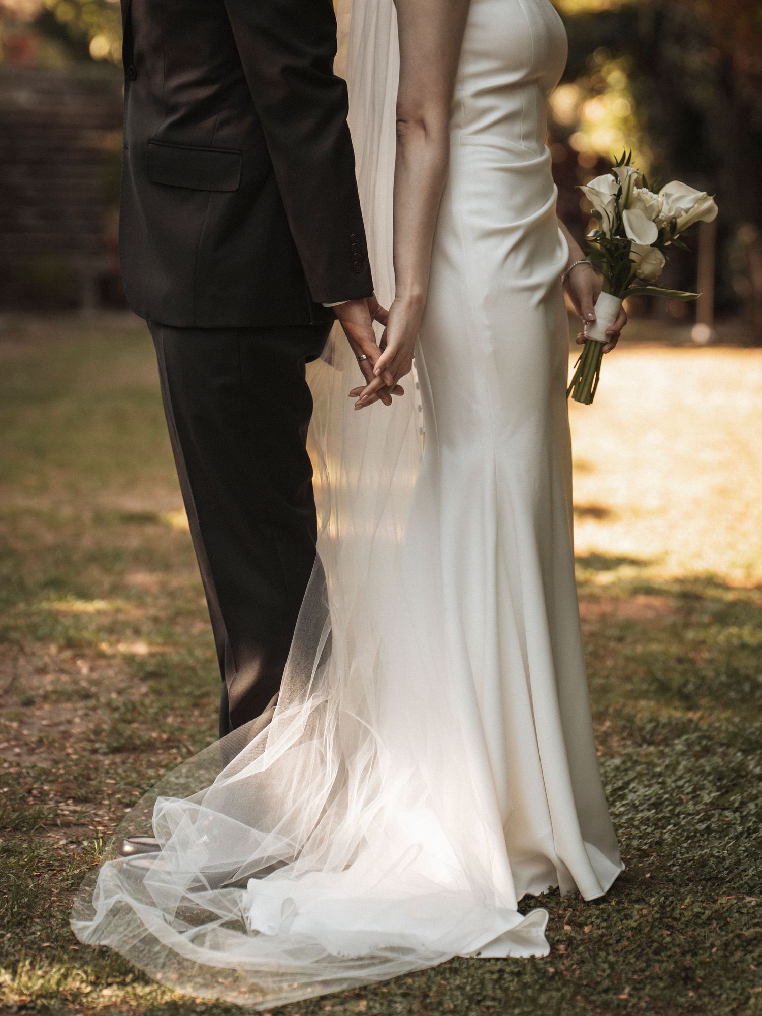 Bride in elegant white gown holding bouquet, holding hands with groom in black suit outdoors.