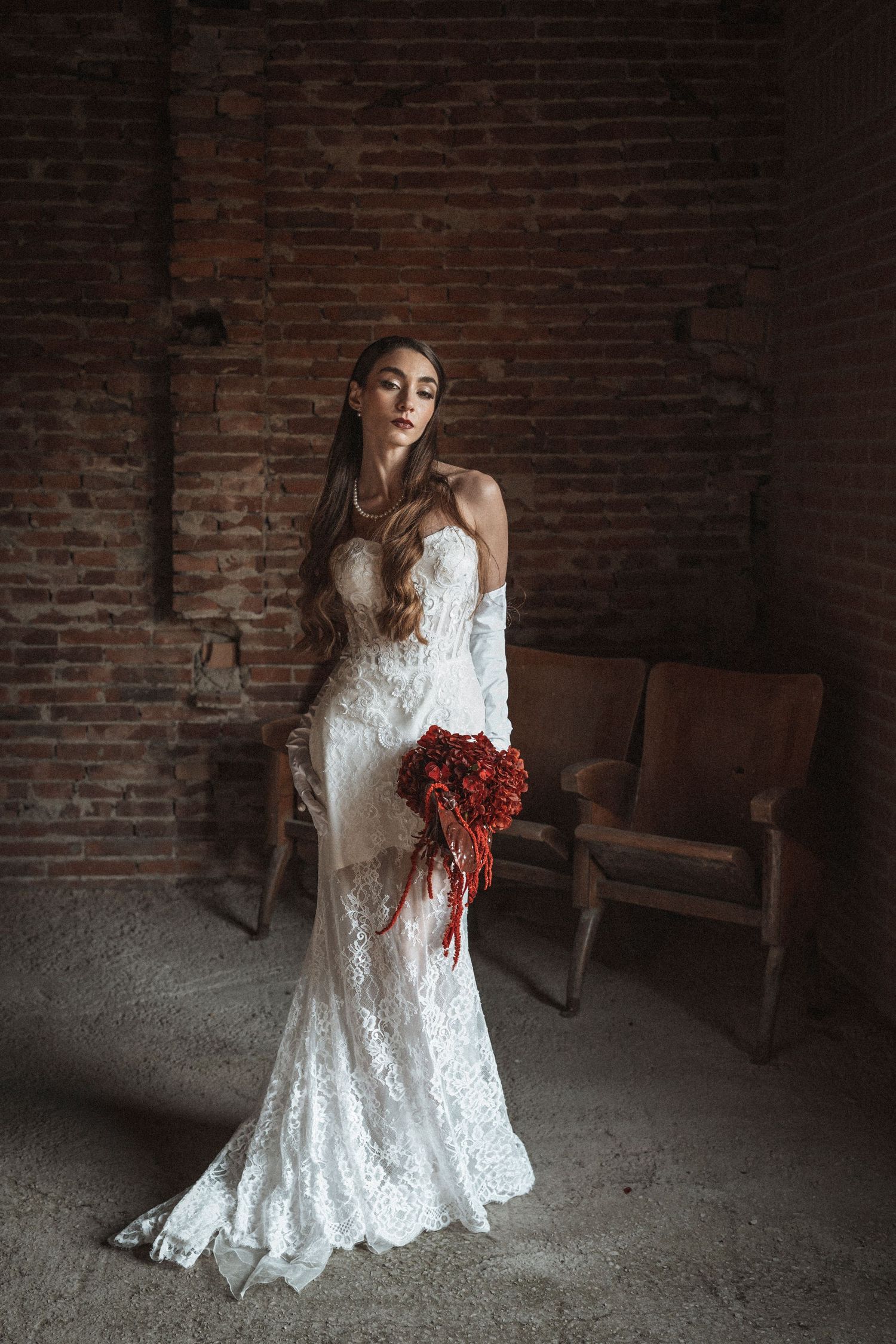 White lace wedding dress photographed in moody lighting against exposed brick wall with red floral accent.