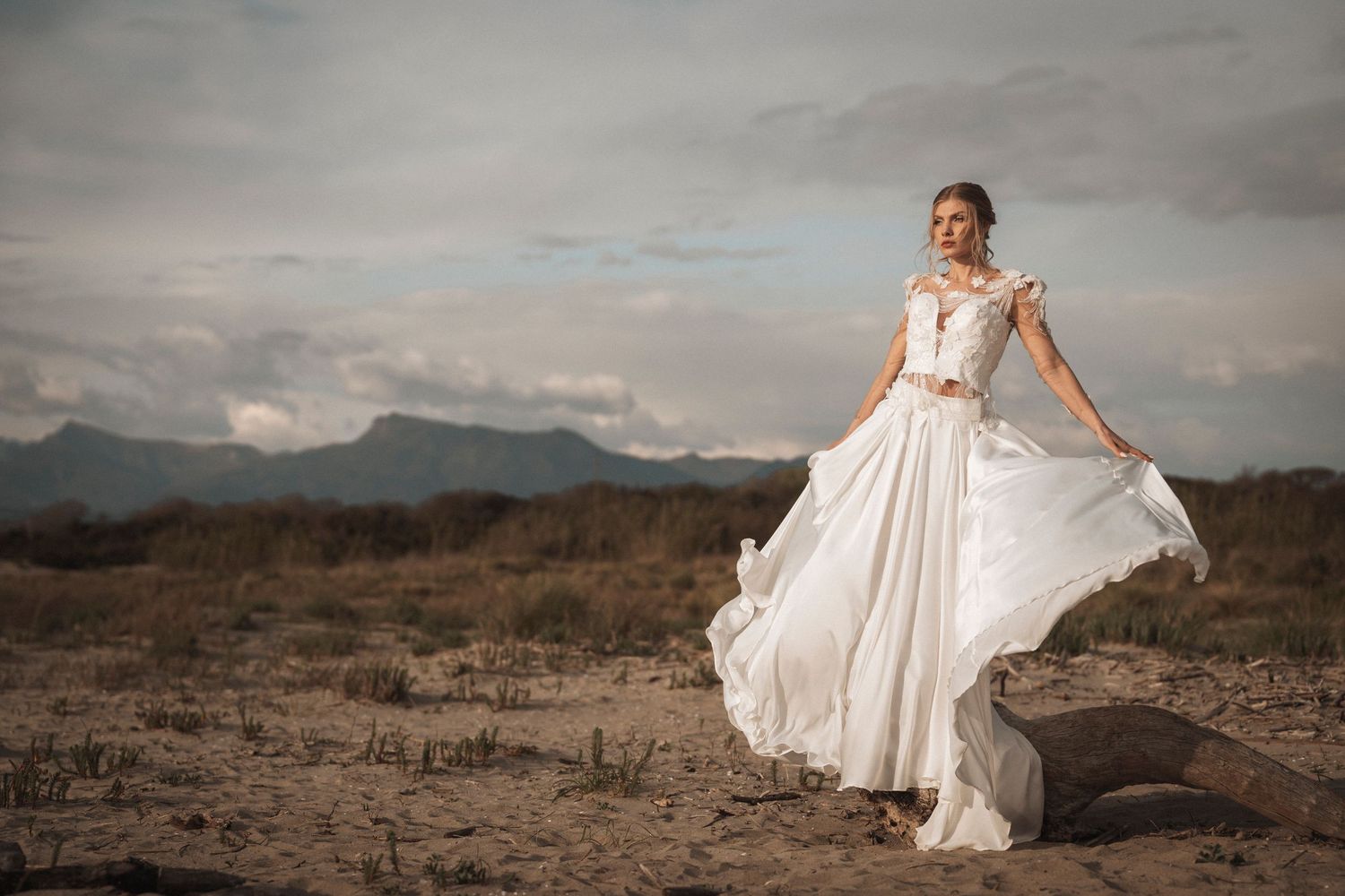 A flowing white wedding dress billows in the wind against a dramatic desert landscape with mountains in the background.