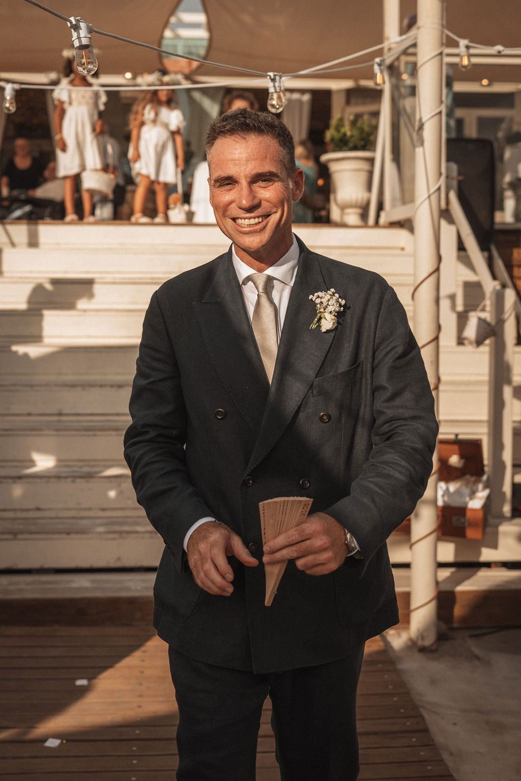 Groomsman in a black suit smiling while holding playing cards on a wooden deck at an outdoor venue.