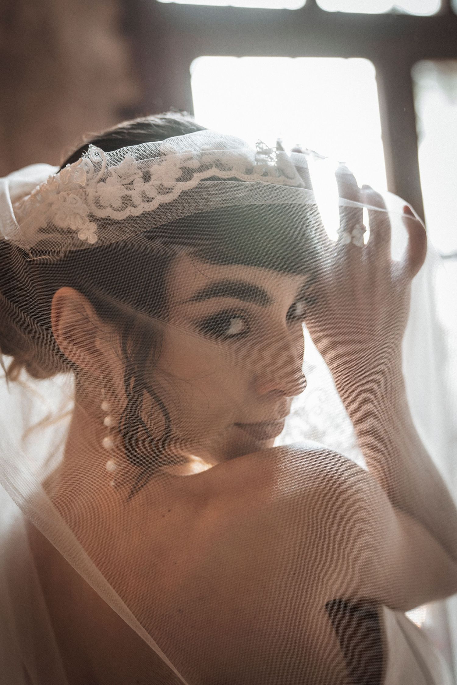 Side profile of bride adjusting lace veil with pearl earrings in romantic natural light.