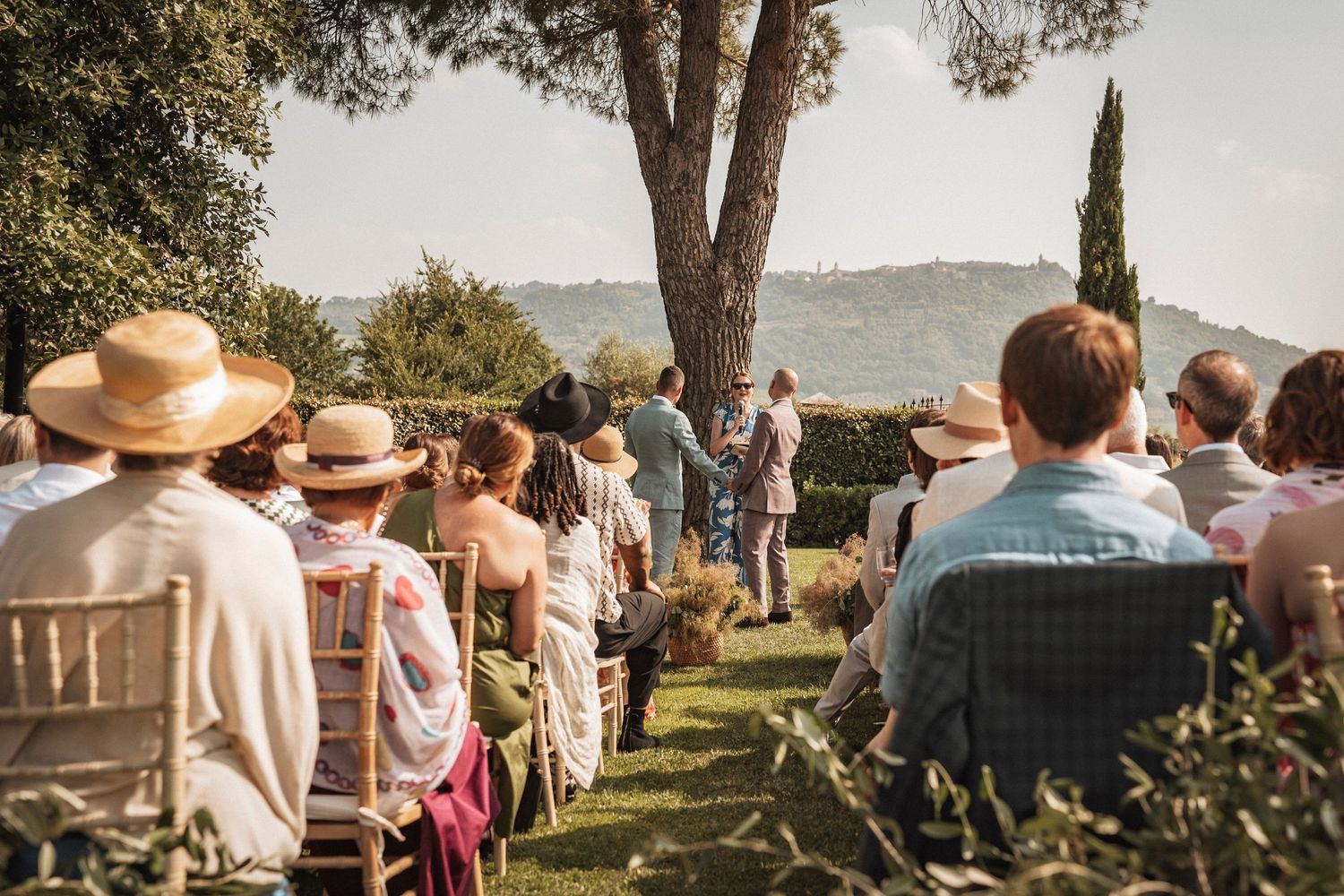 Panoramic view of an outdoor wedding ceremony with guests seated beneath tall trees.