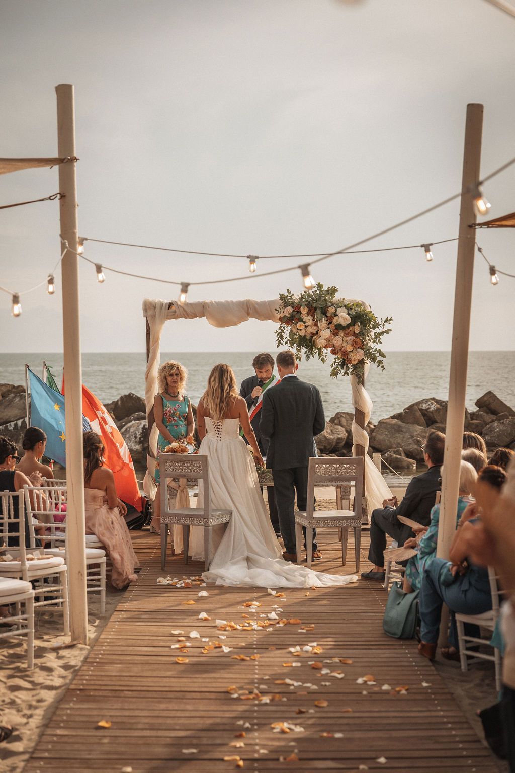 Wedding guests seated on a wooden deck by the ocean with string lights and rose petals scattered.
