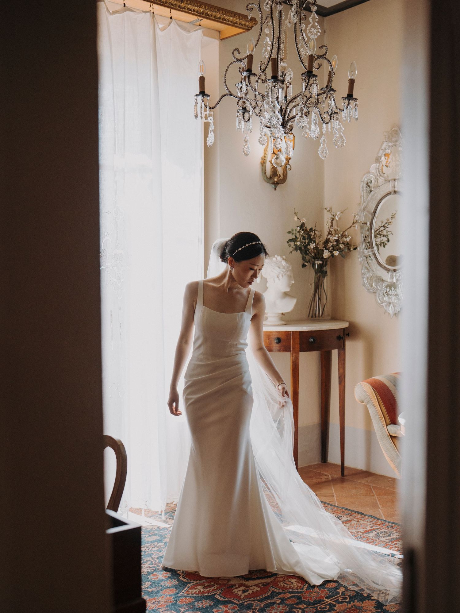 Bride in elegant white satin mermaid gown with train stands in ornate room with crystal chandelier.