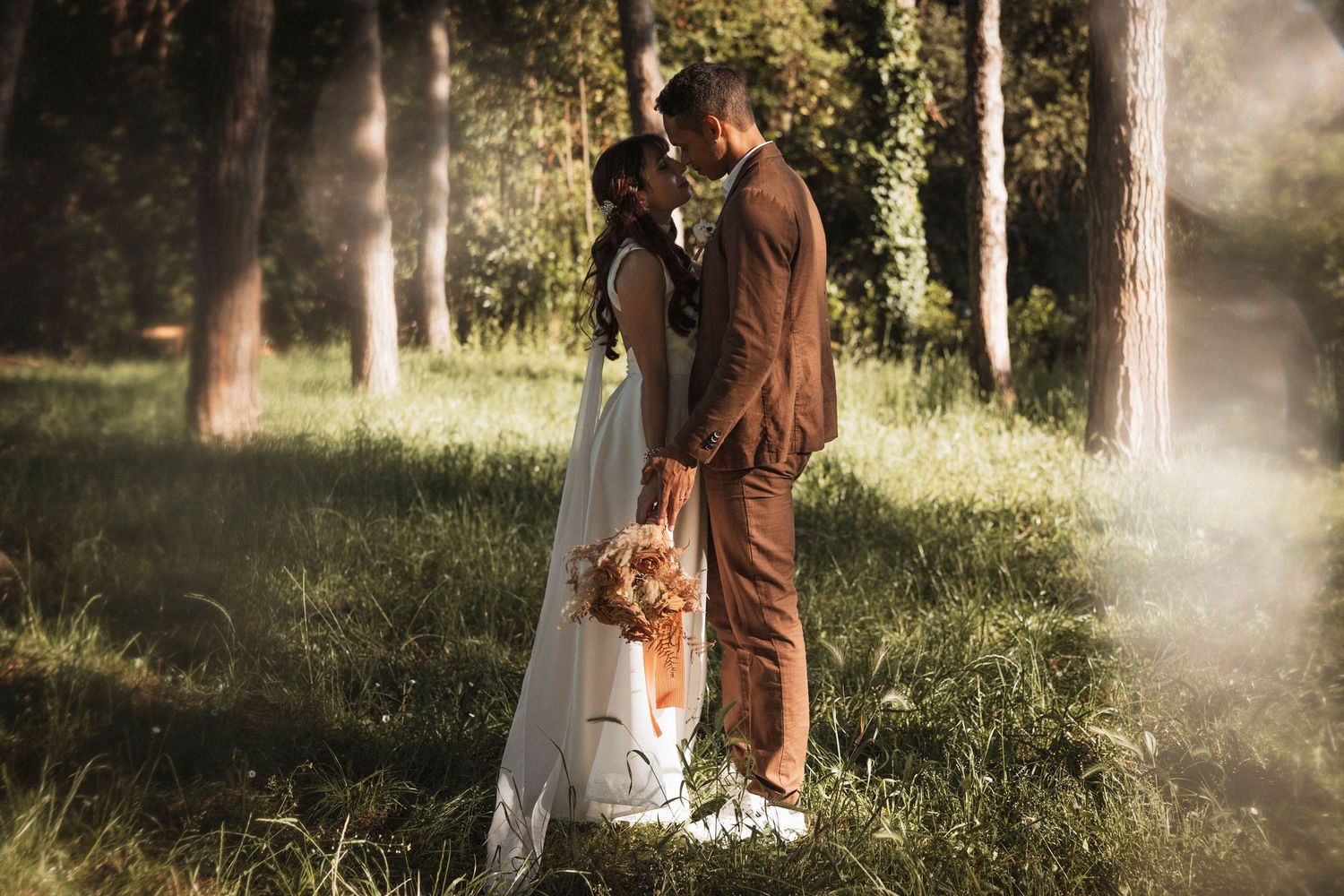 A couple in brown and white outfits poses together in a misty forest setting during a romantic photoshoot.