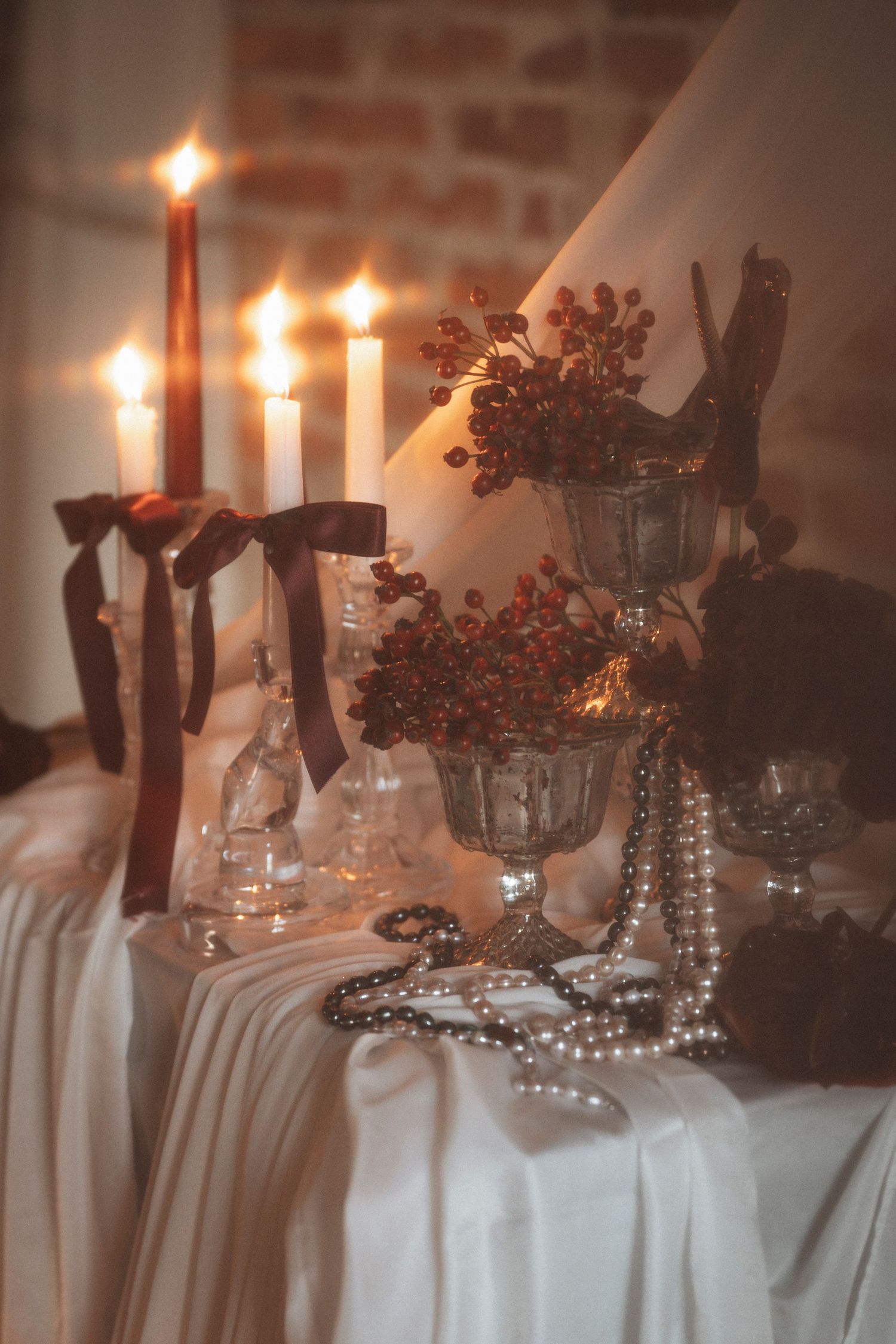 Romantic candlelit table setting with pearls and burgundy florals against rustic brick walls.