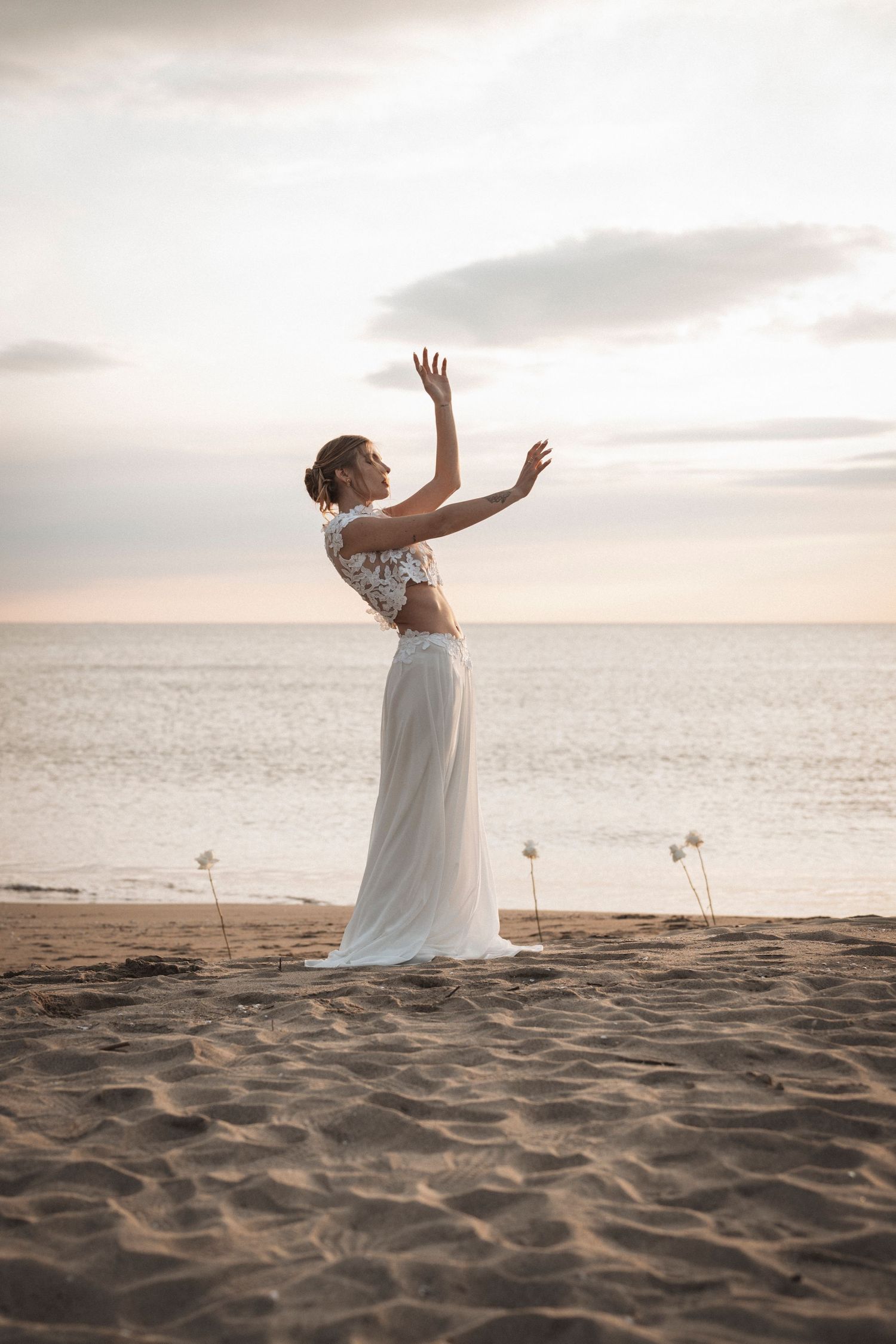 A figure in a flowing white dress dances joyfully on a sandy beach at sunset with arms raised to the sky.