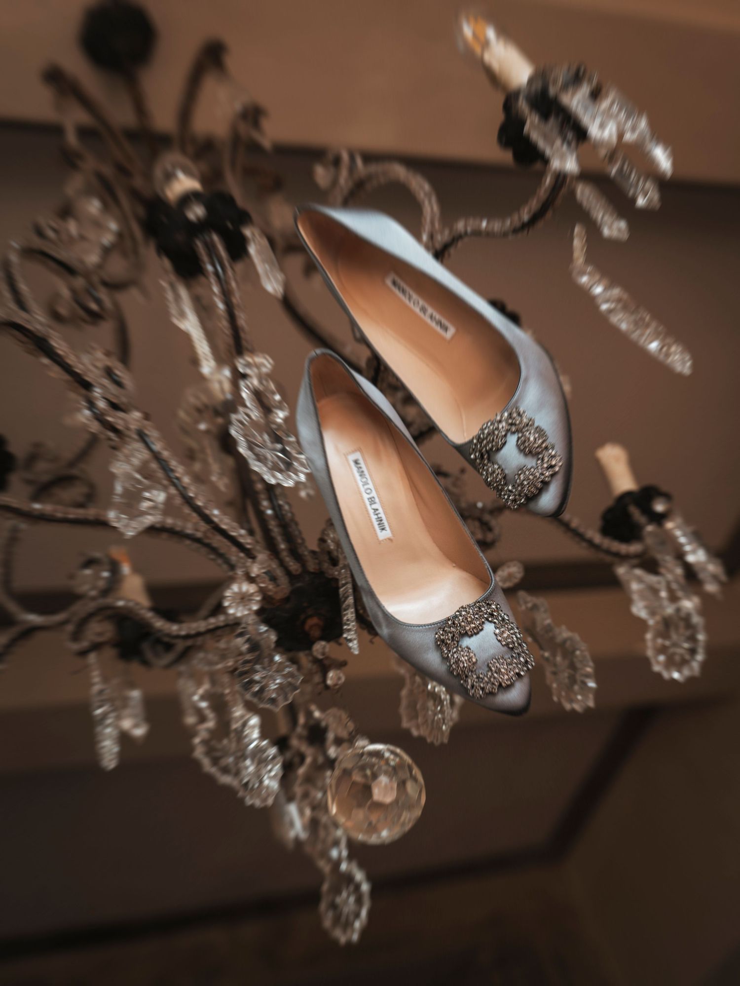 Light blue Manolo Blahnik crystal-buckle heels draped elegantly over an ornate silver chandelier.
