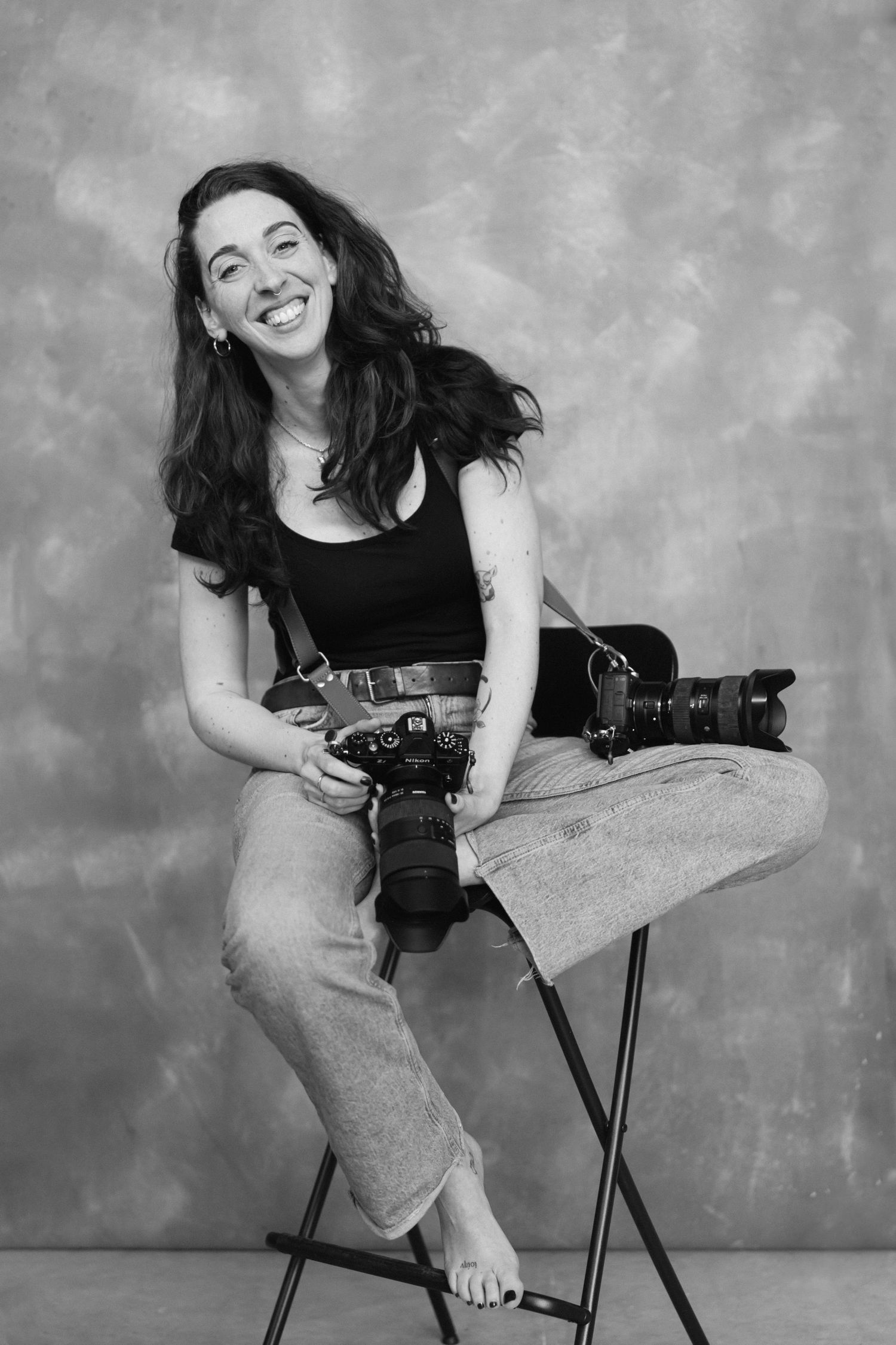 Black and white portrait of a smiling woman sitting on a stool holding two cameras in a photography studio.