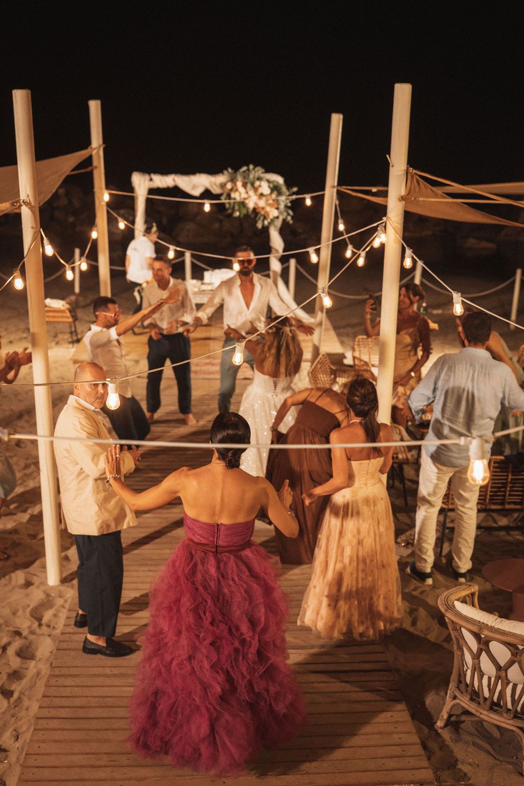Guests gather on a wooden deck decorated with string lights and fabric drapes for an evening celebration.