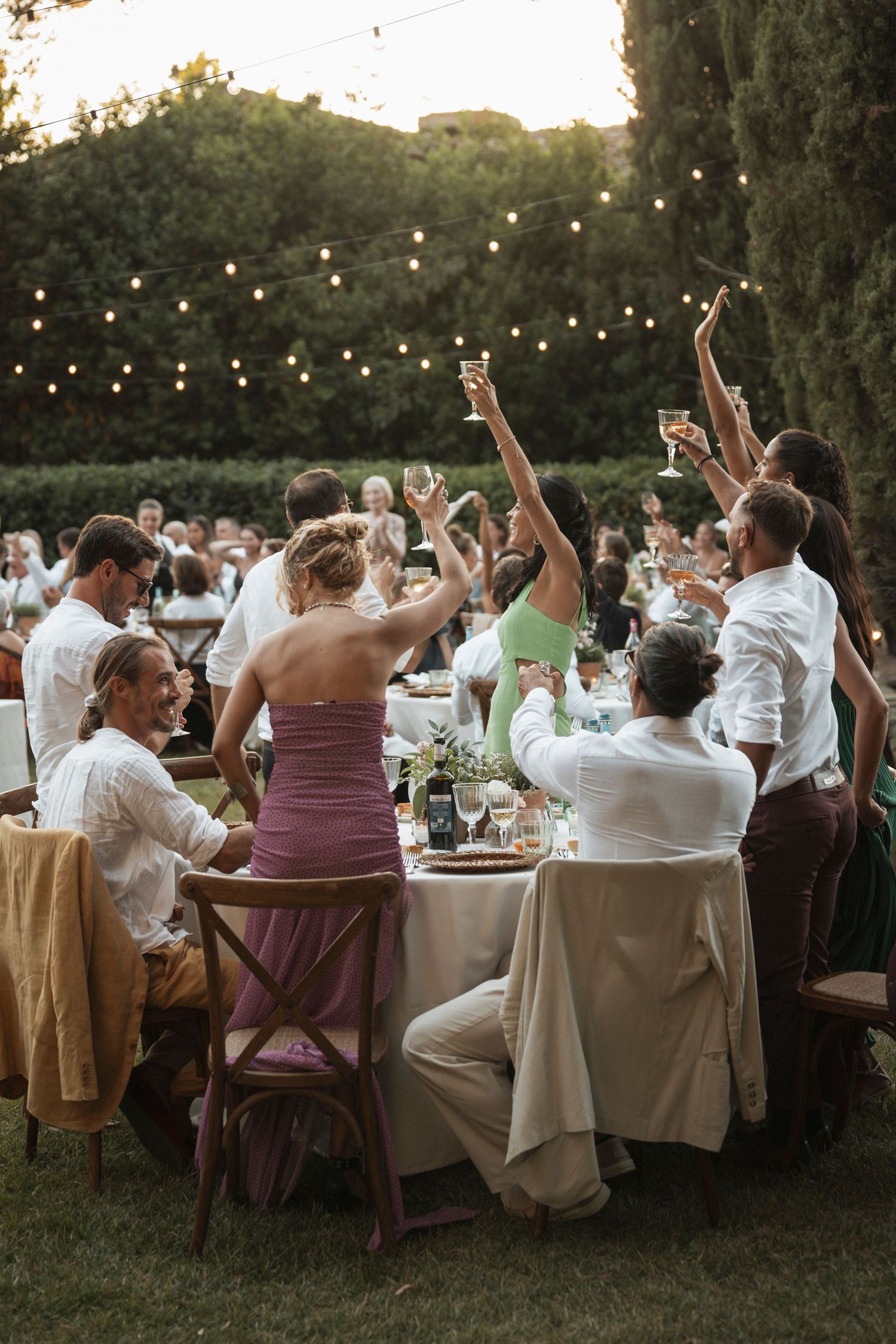 Wedding reception guests raising their glasses in a toast under string lights at sunset.