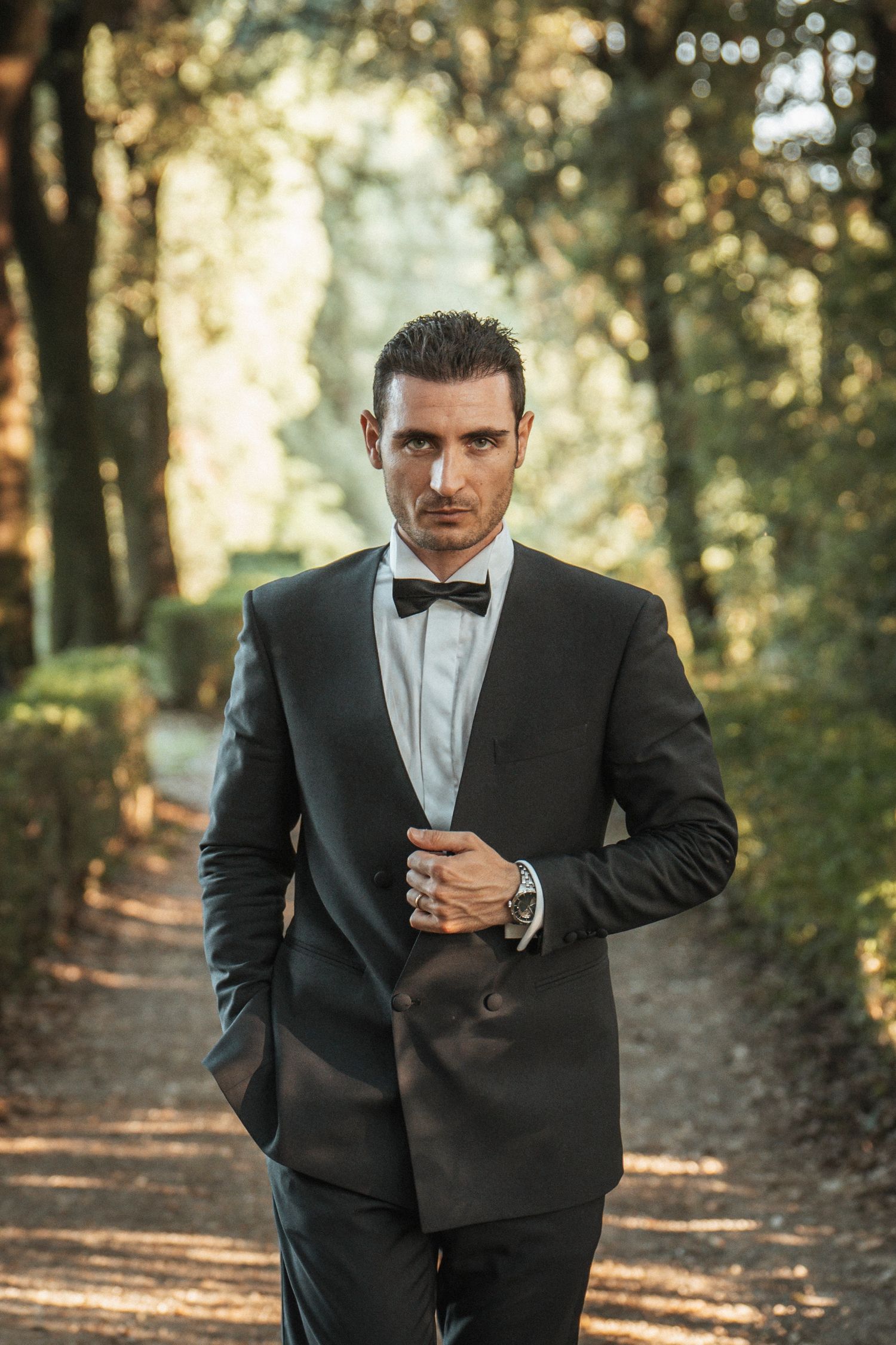 Handsome groom in black tuxedo with bow tie stands confidently on a tree-lined garden path.