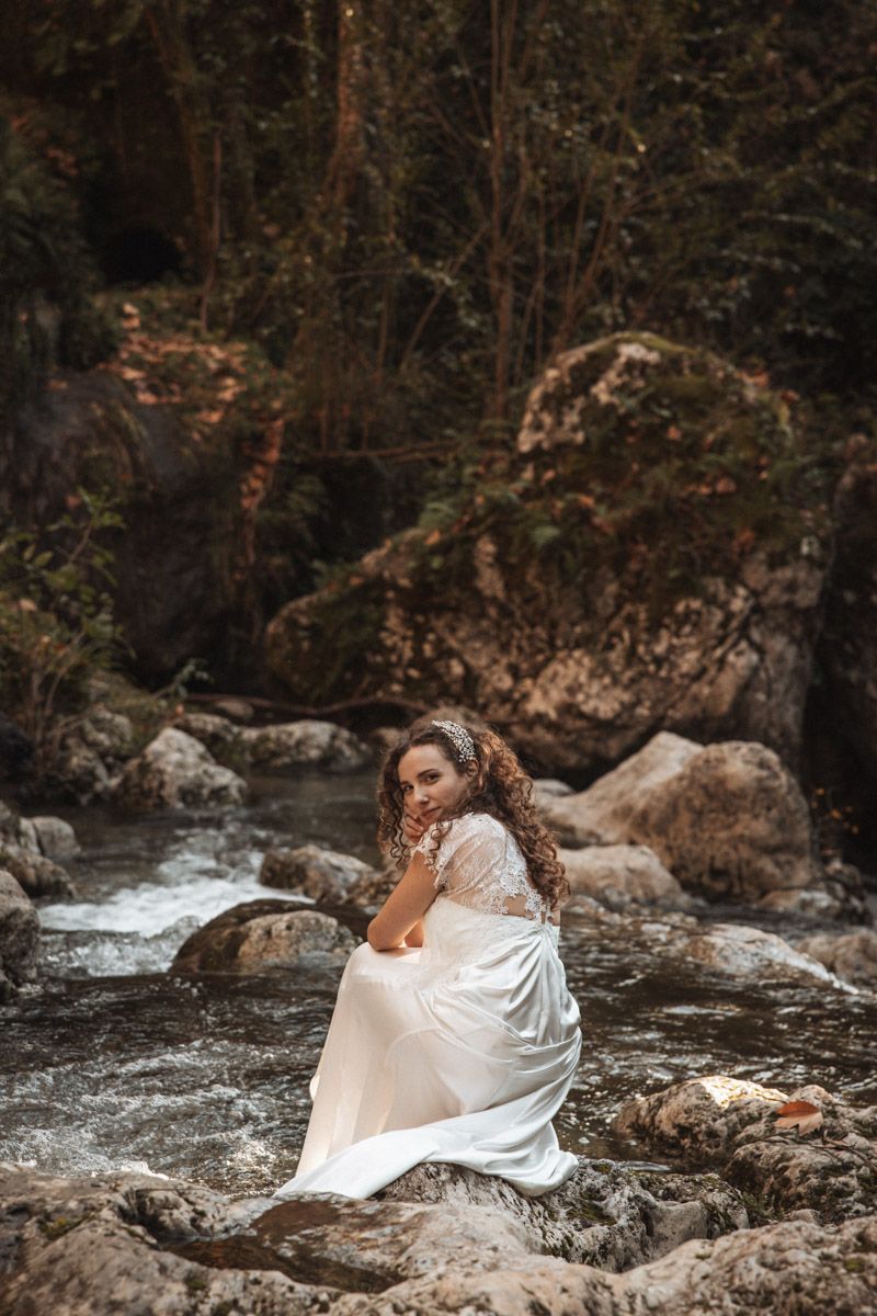 A flowing white dress drapes over rocky terrain beside a rushing stream in a moody forest setting.