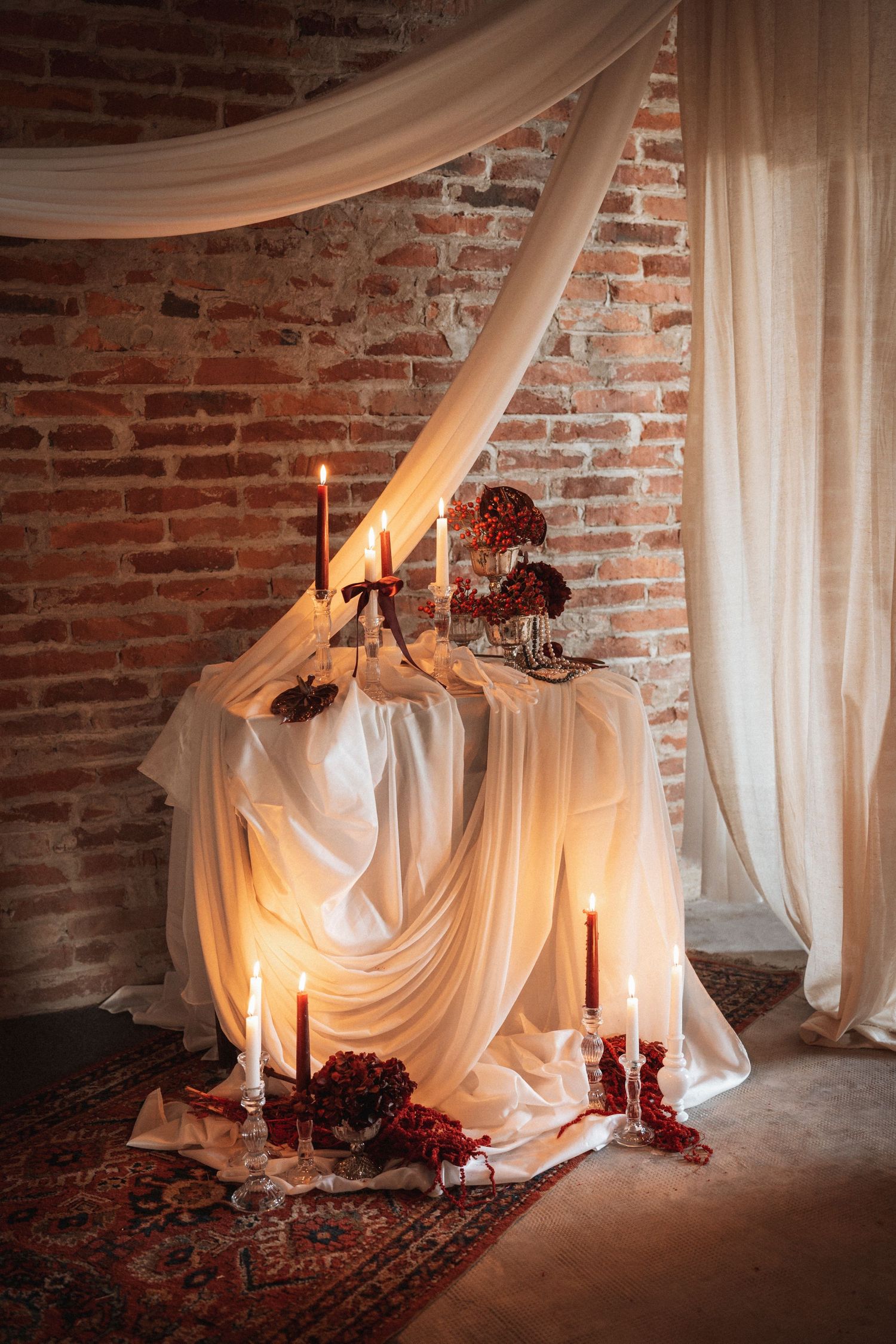 Romantic wedding setup with draped white fabric, red flowers, and candles against exposed brick wall.