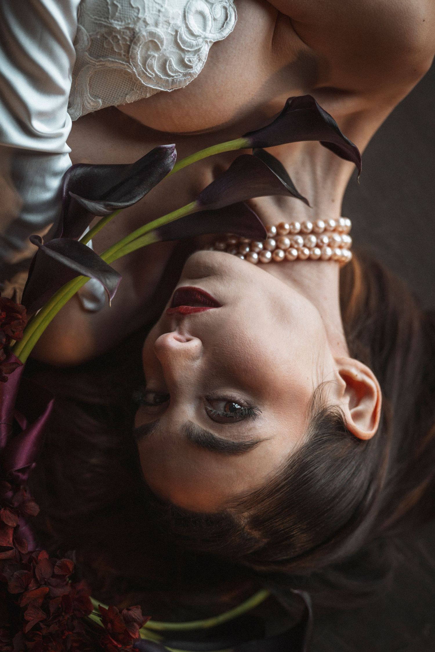 Artistic portrait featuring pearl necklace and dark lipstick in moody lighting with calla lilies.