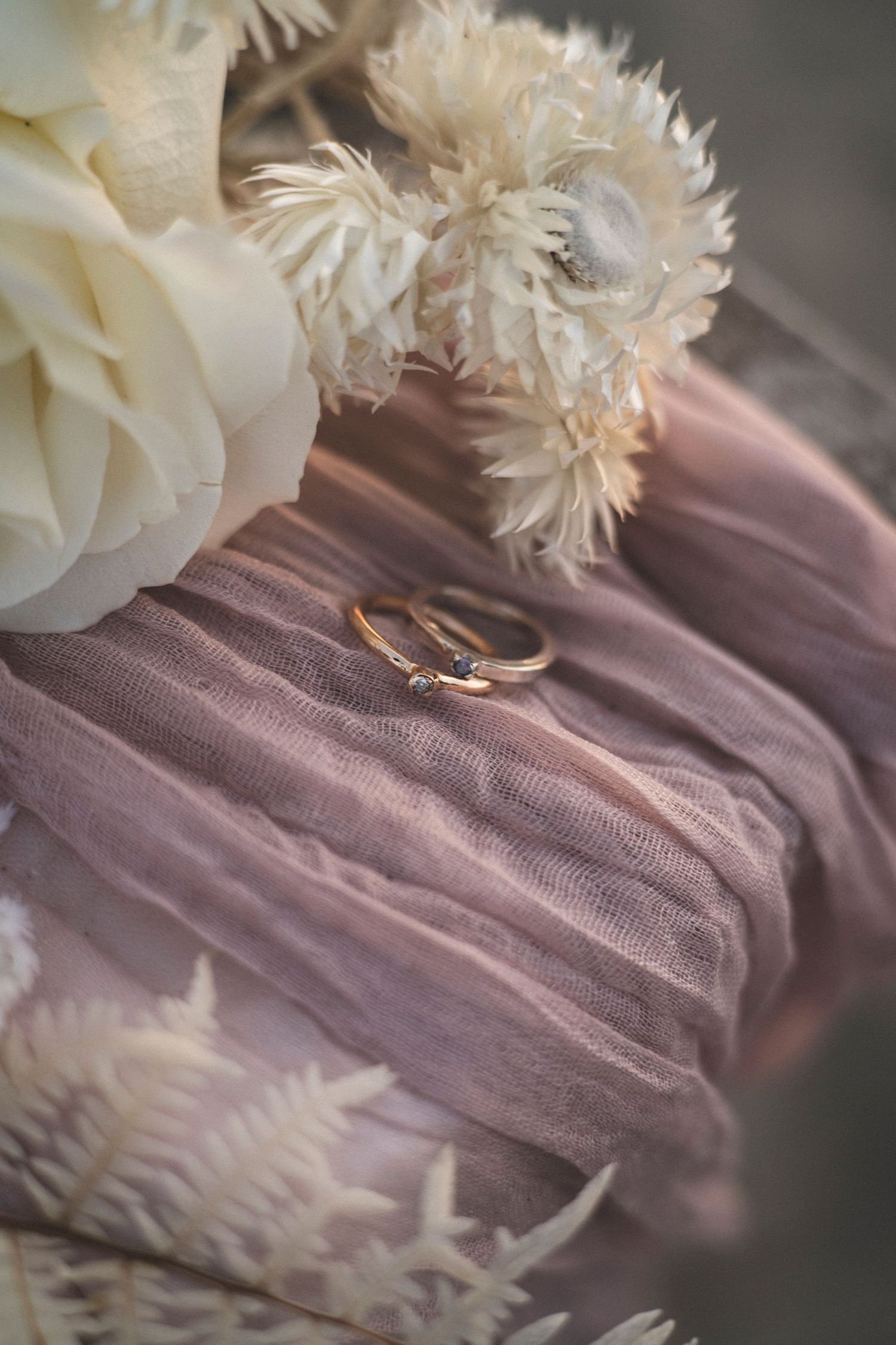 Wedding rings rest on dusty pink tulle fabric surrounded by delicate white flowers in a romantic arrangement.