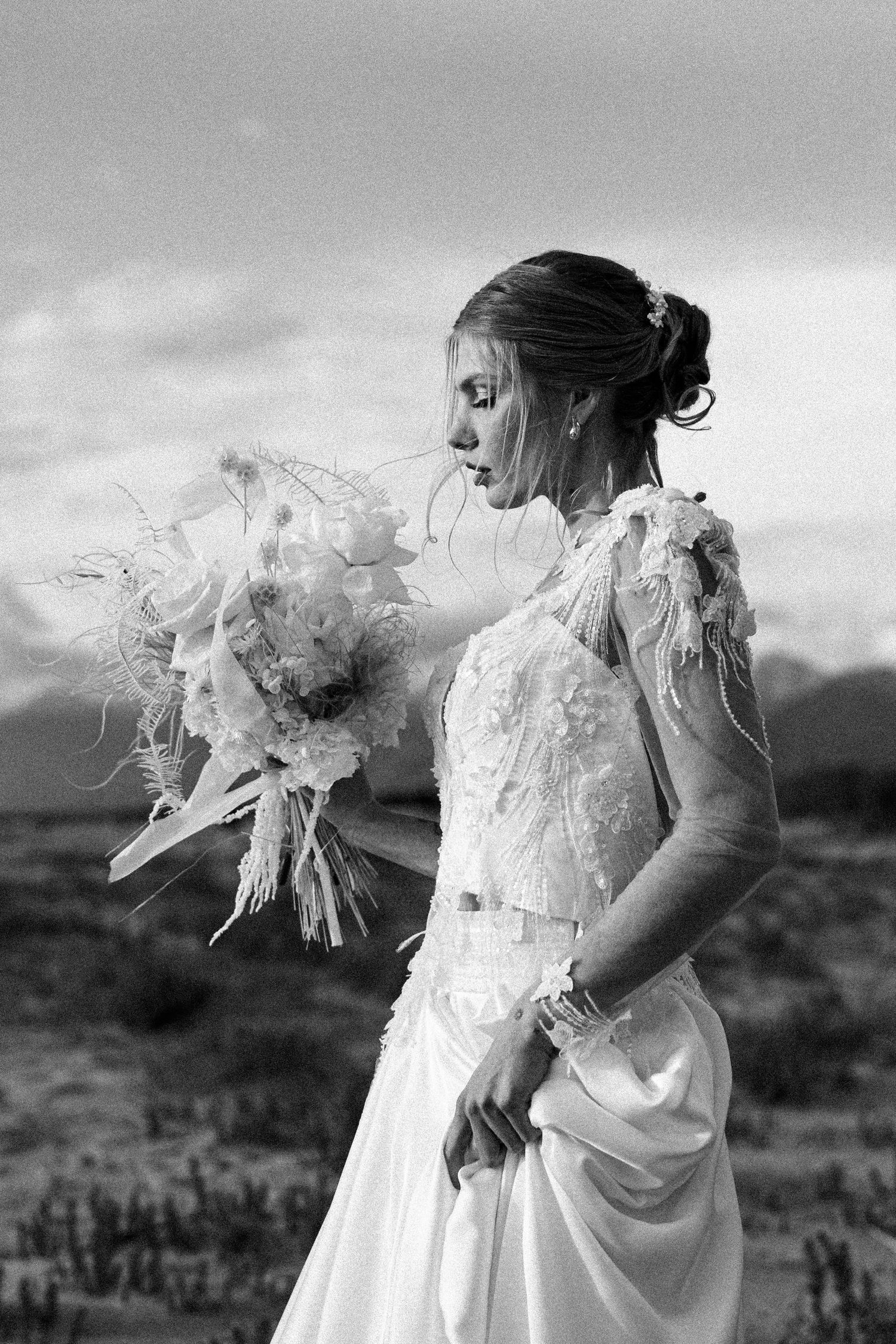 A black and white bridal portrait showcasing an elegant lace wedding gown and bouquet in a desert setting.