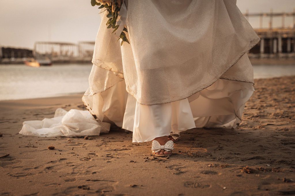 Close-up details of a white wedding dress flowing across sandy beach.