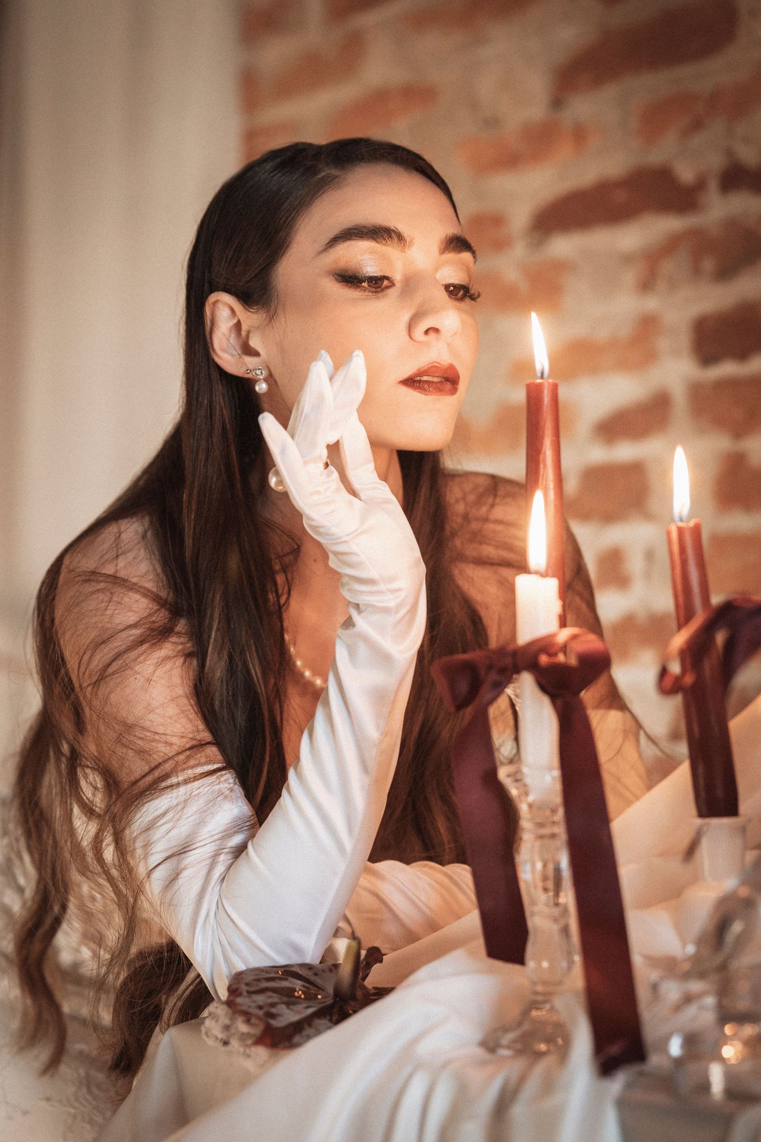 Atmospheric portrait with candlelight featuring white gloves and red lipstick against a brick wall backdrop.