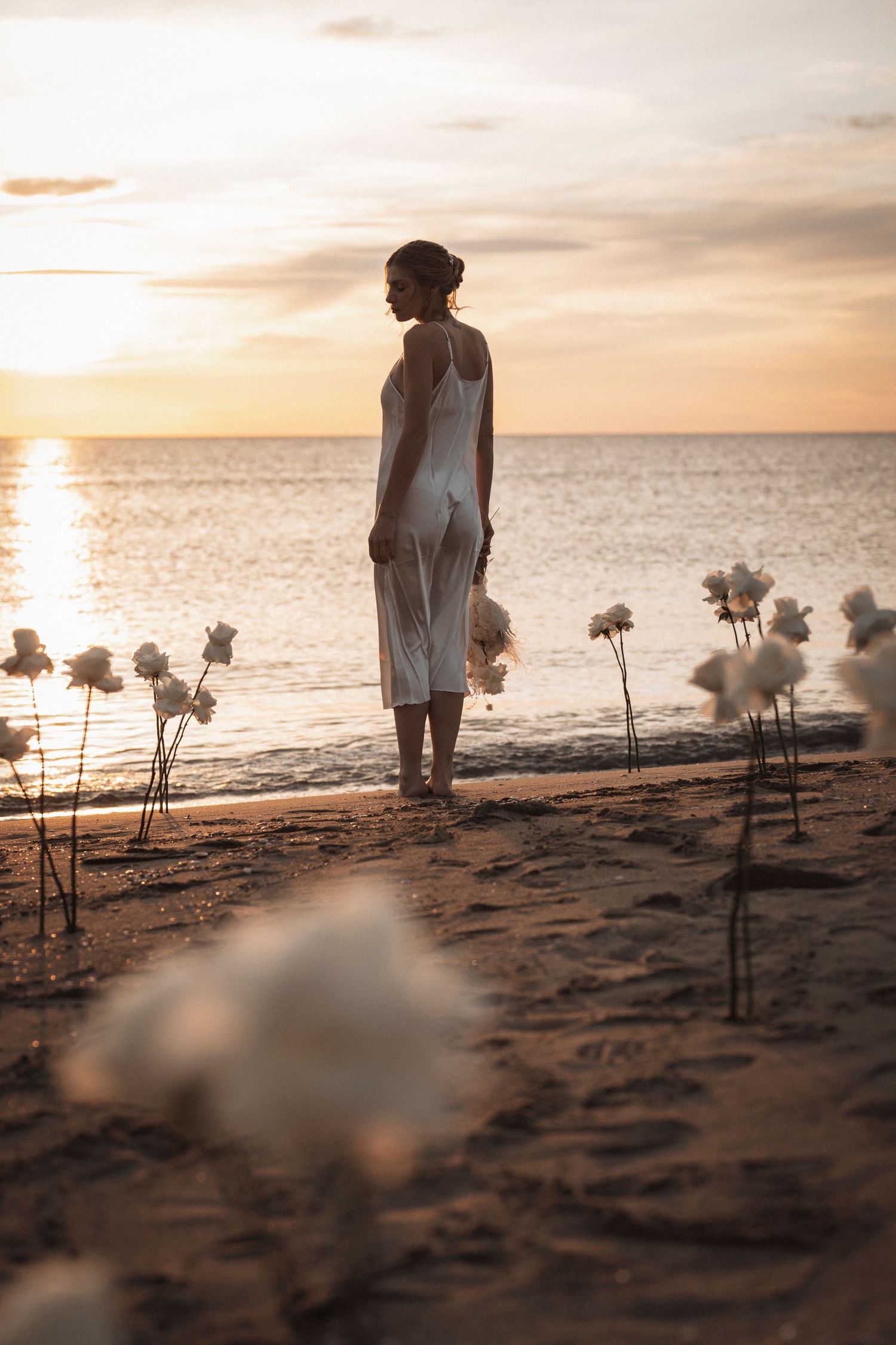 A silhouette of a figure in white stands on a beach at sunset with flowers planted in the sand along the shoreline.