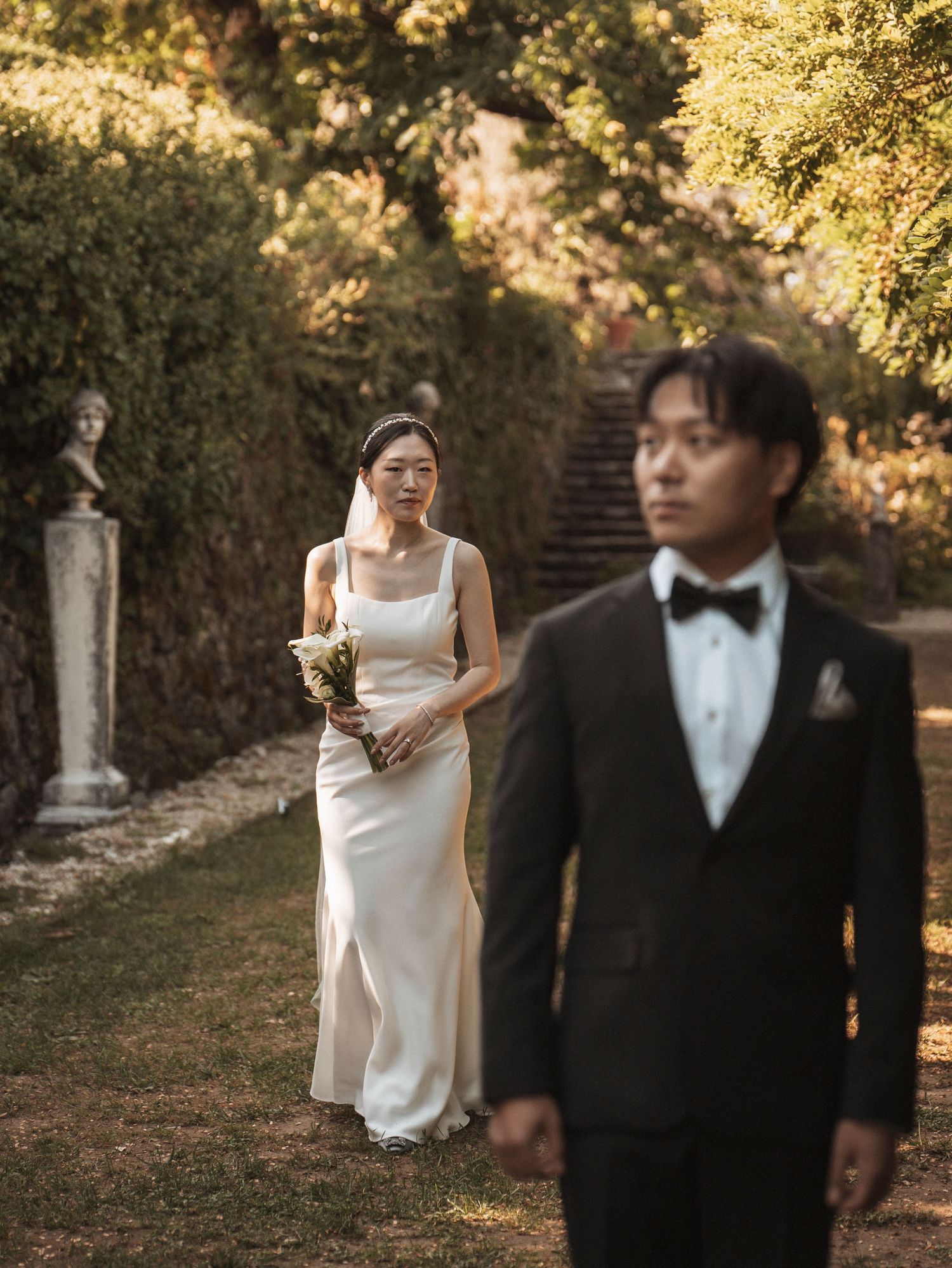 Bride in white gown with bouquet walks toward groom in black tuxedo in a lush garden wedding setting.