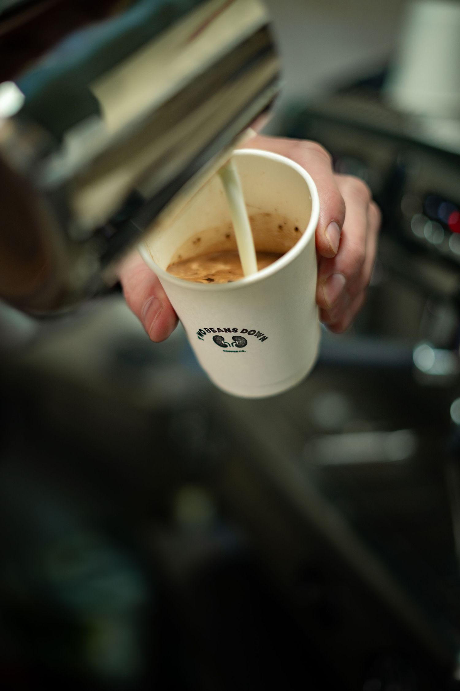 Barista pouring steamed milk into a branded Two Beans Down paper coffee cup at espresso machine.