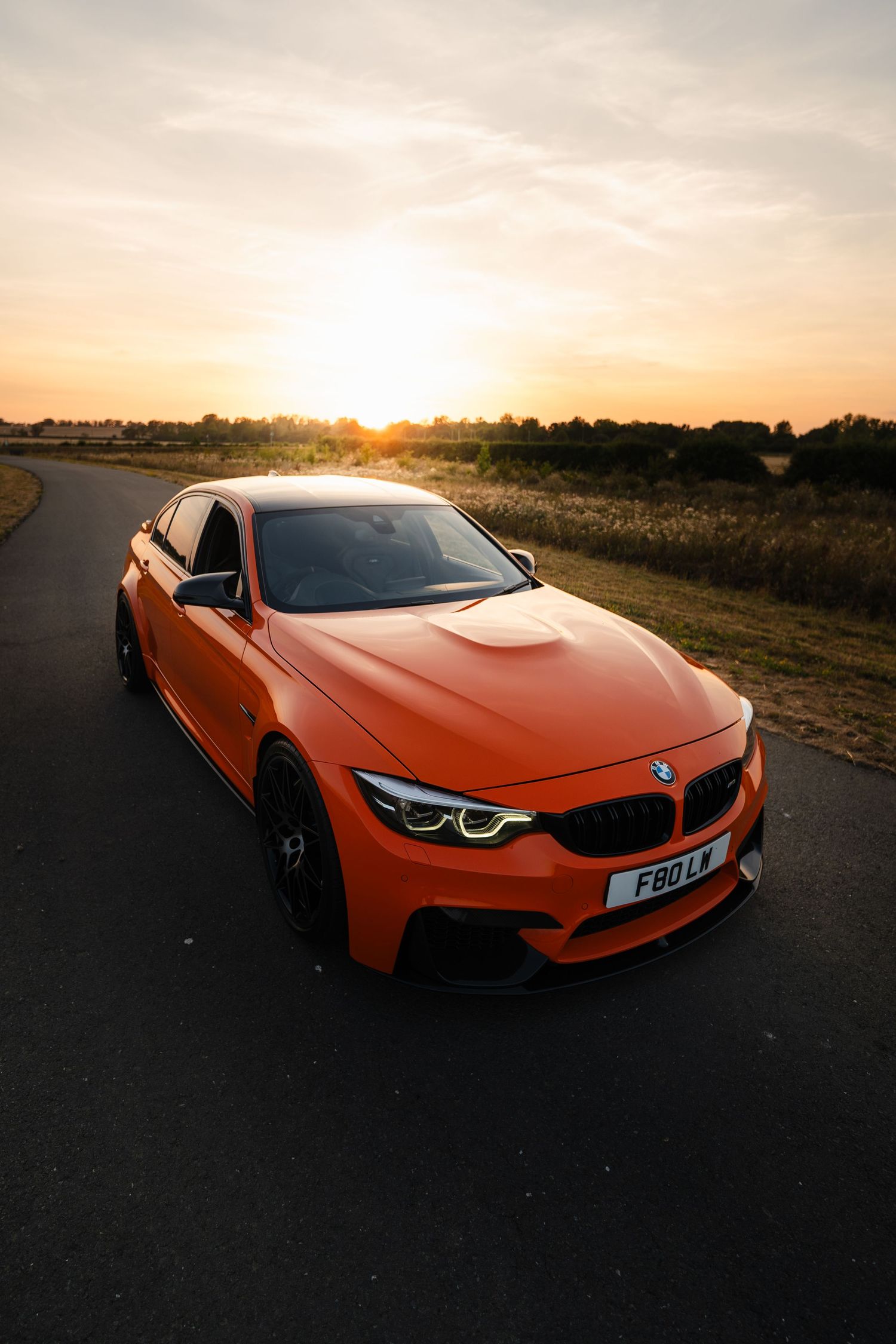 Orange BMW M4 sports car parked on a road at sunset with golden hour lighting and rural landscape background.