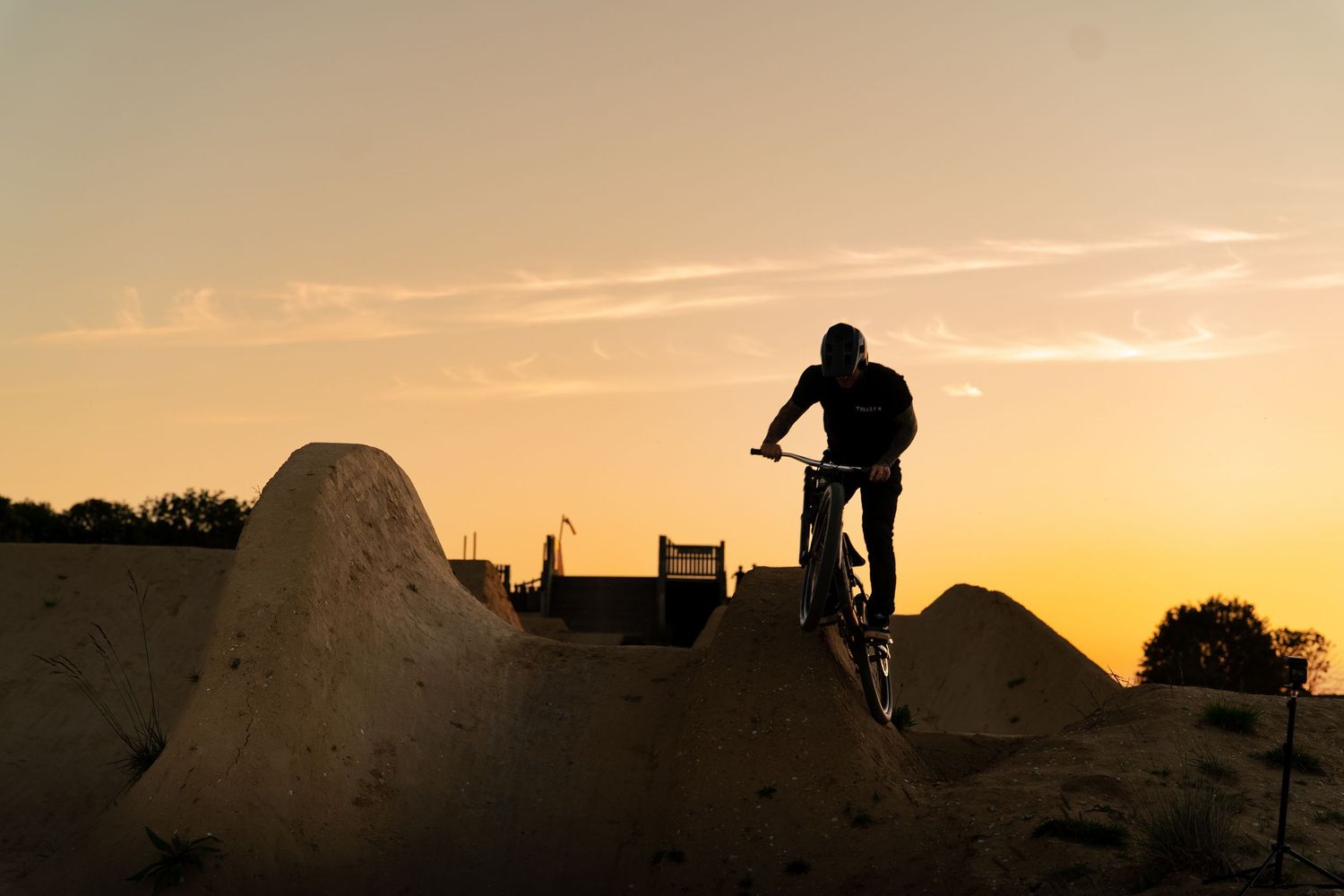 Silhouette of a cyclist on a dirt jump ramp at an outdoor bike park during sunset.