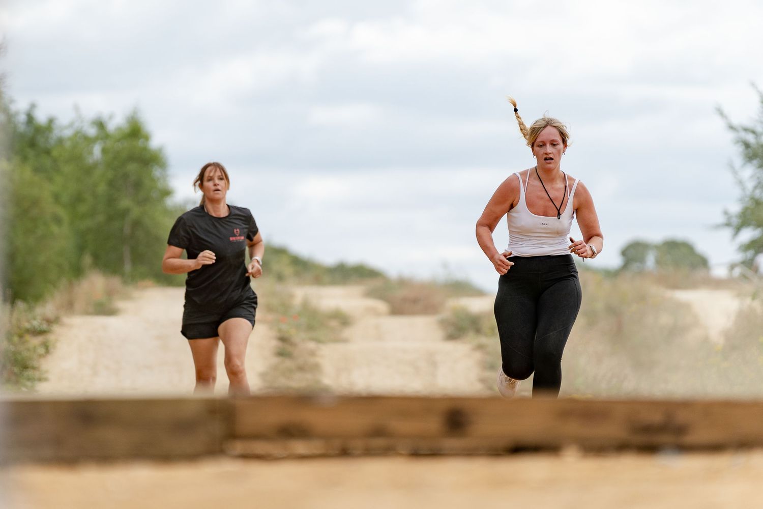 Two women running outdoors on a sandy path with trees in the background during a race or training exercise.