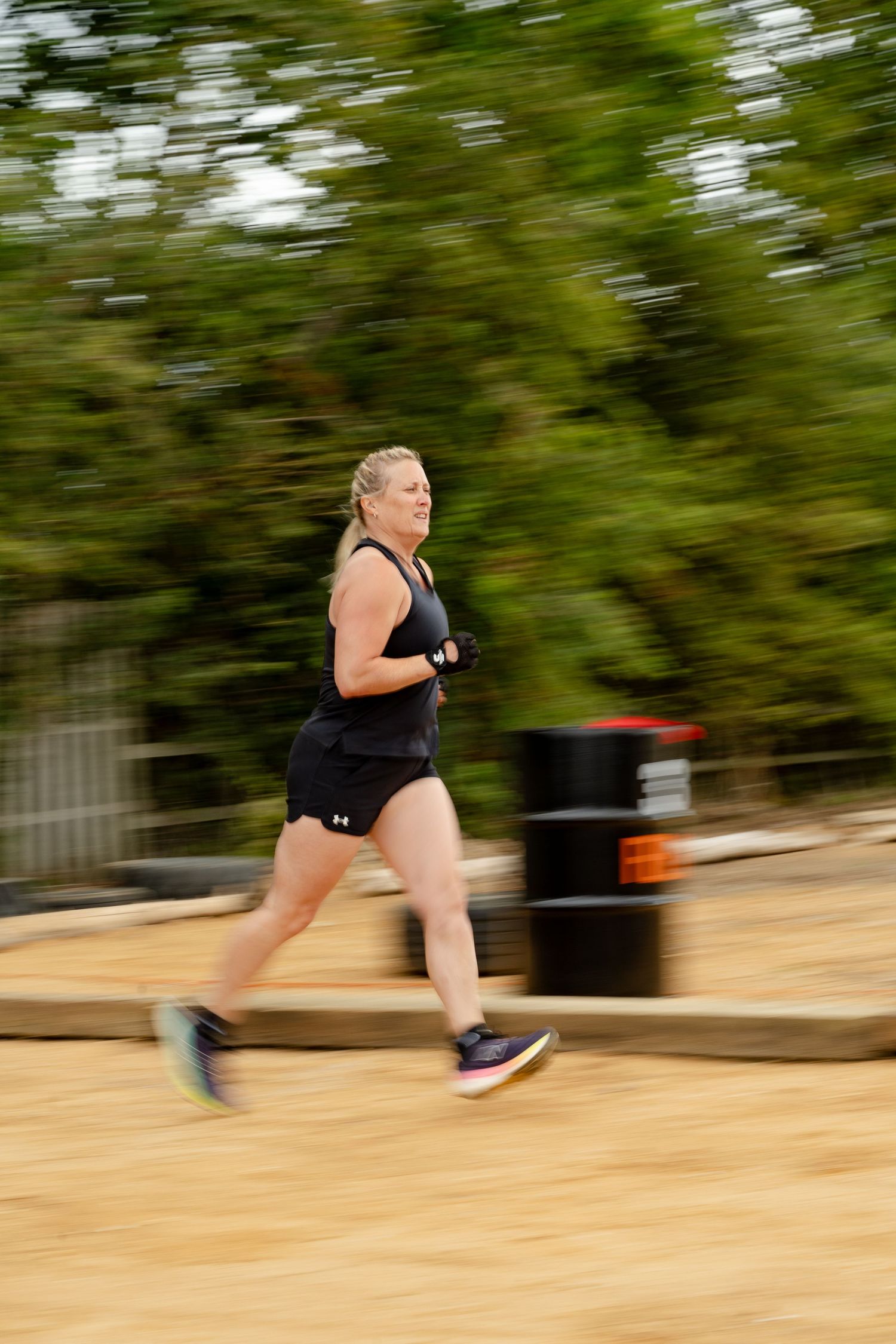 Woman in black athletic wear running outdoors on a dirt path with motion blur effect and green trees in background.