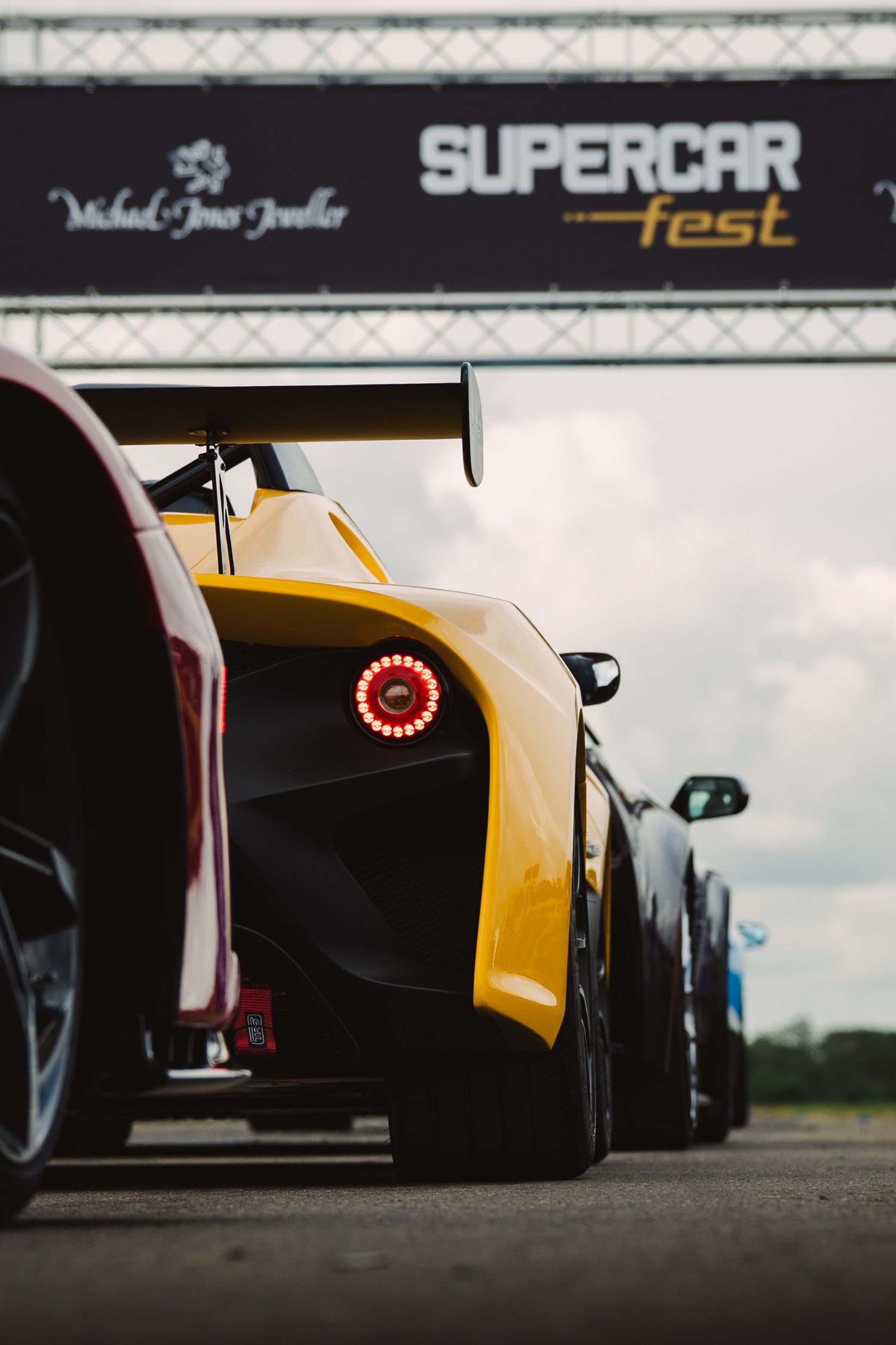 Yellow supercar at Supercar Fest event lined up on track with other exotic cars under branded gantry.