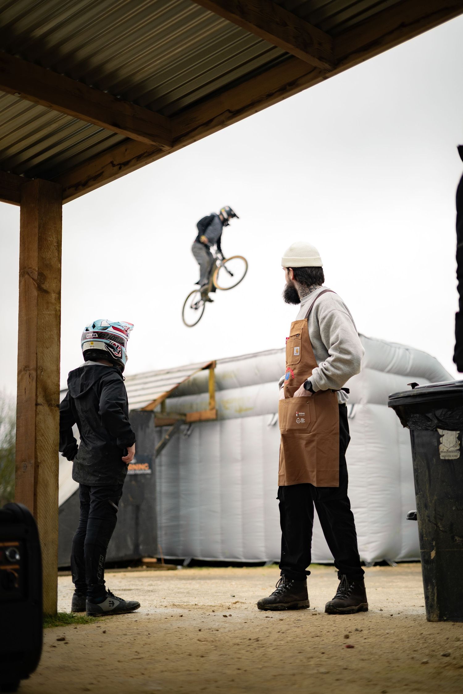 A bearded barista and young BMX rider watch a cyclist perform a jump trick at an outdoor skate park.