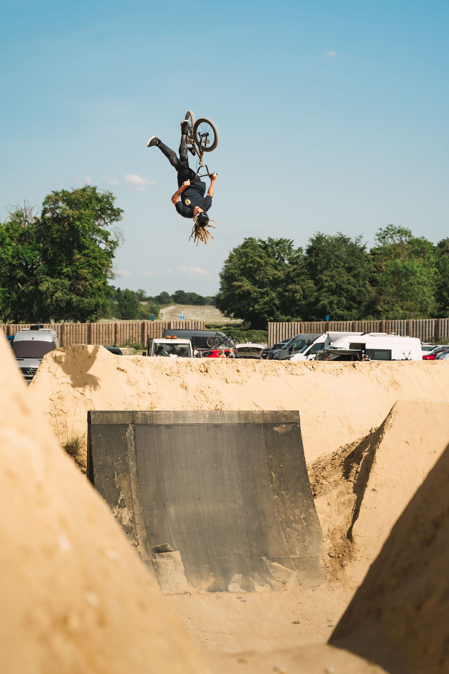 BMX rider executing a dramatic backflip over a metal ramp at a sandy outdoor dirt jump park.