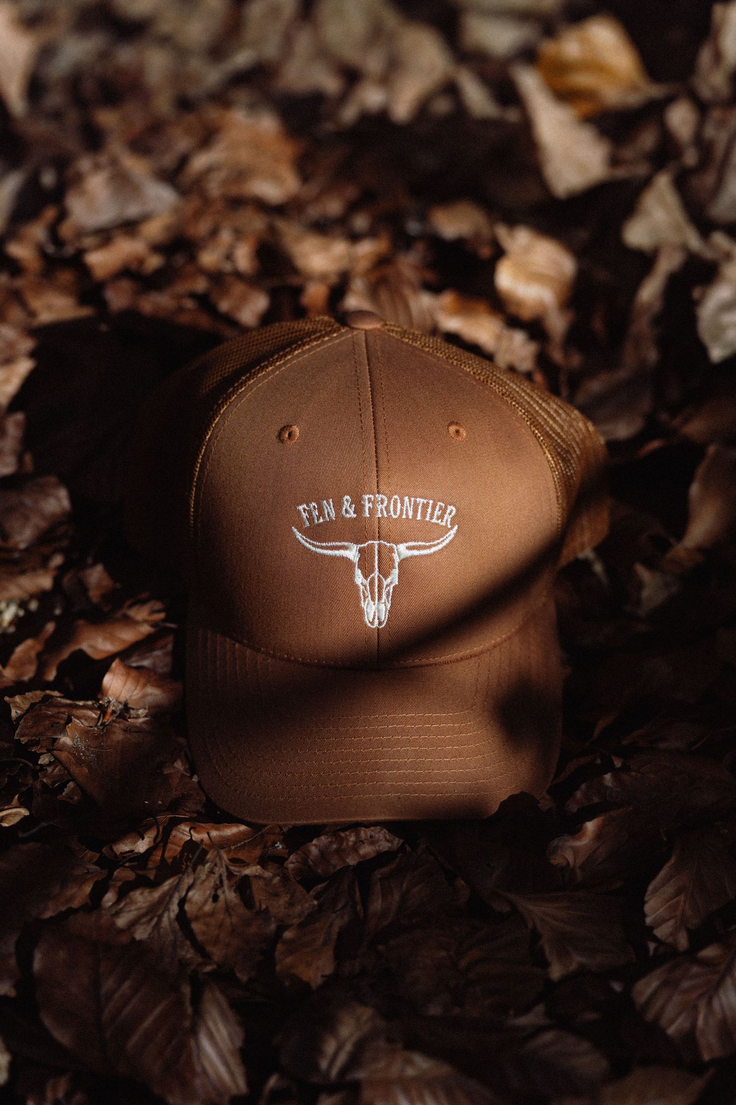 Brown Fen & Frontier trucker hat with longhorn skull logo resting on autumn leaves.