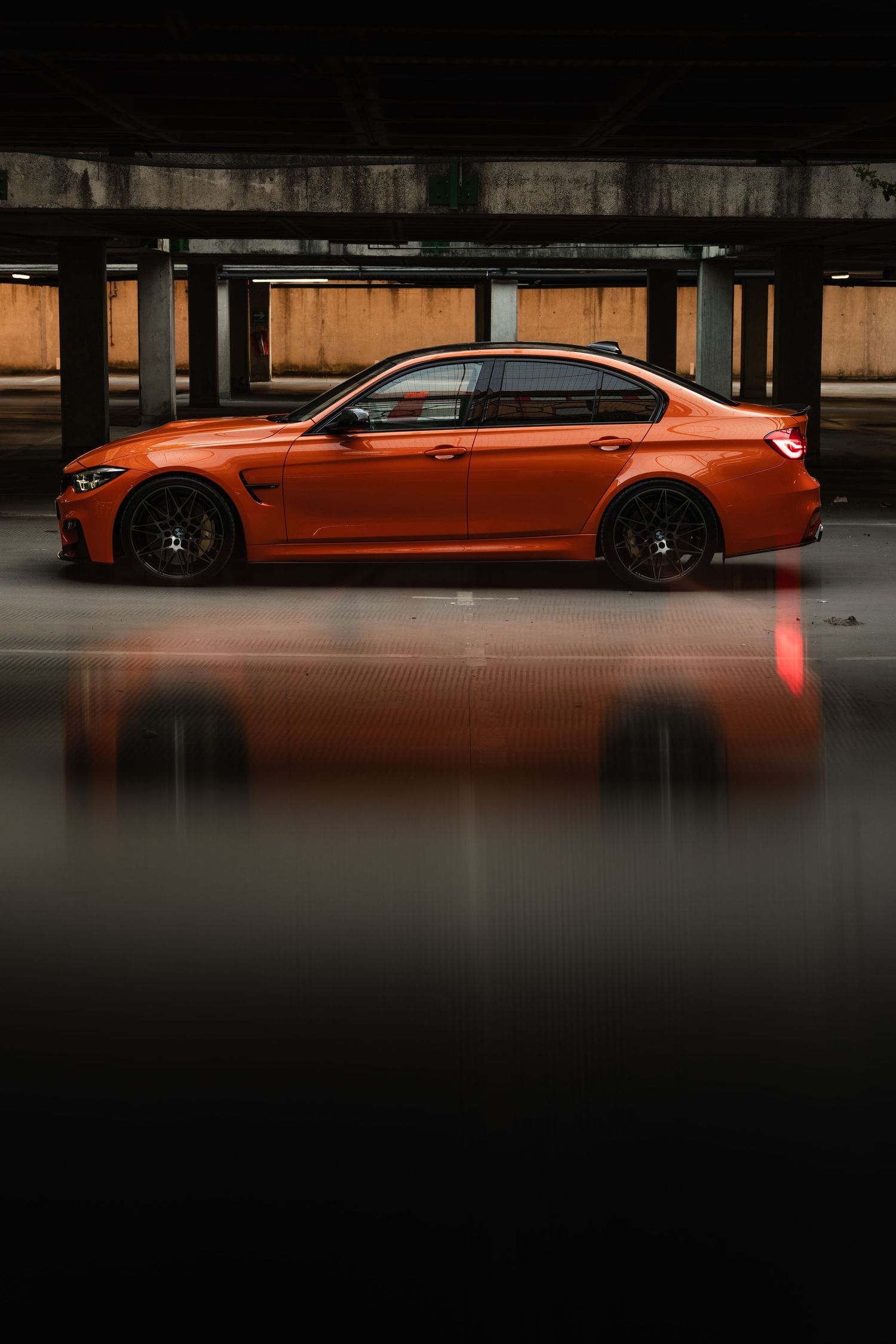 Orange BMW M3 sedan parked in a dark garage, showcasing its sleek profile and black wheels on a reflective floor.