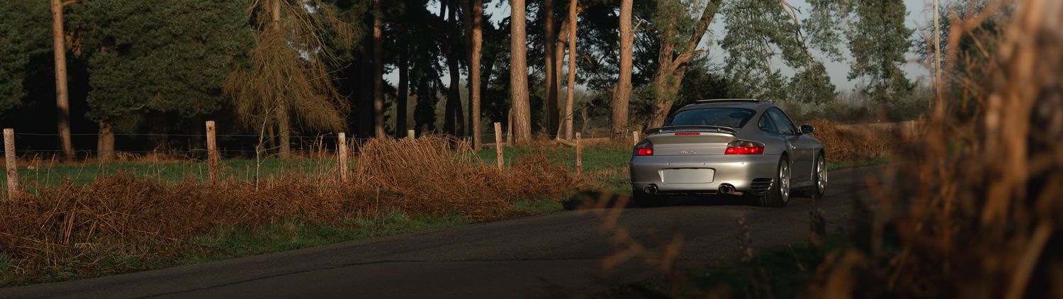 Silver Porsche 911 Turbo driving along a rural tree-lined road in autumn landscape.