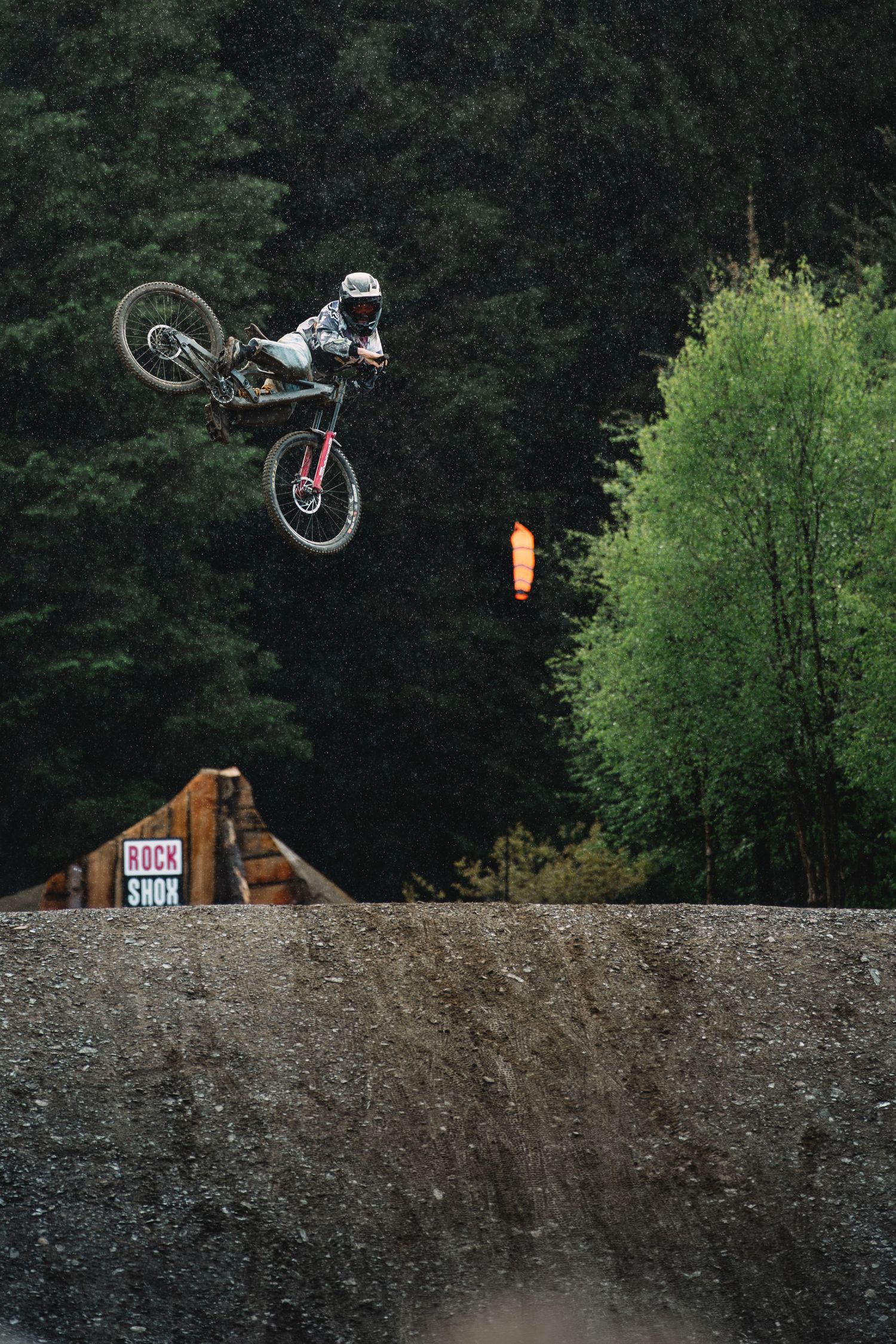 A mountain biker performs a high aerial trick off a dirt jump ramp with dense forest trees in the background.