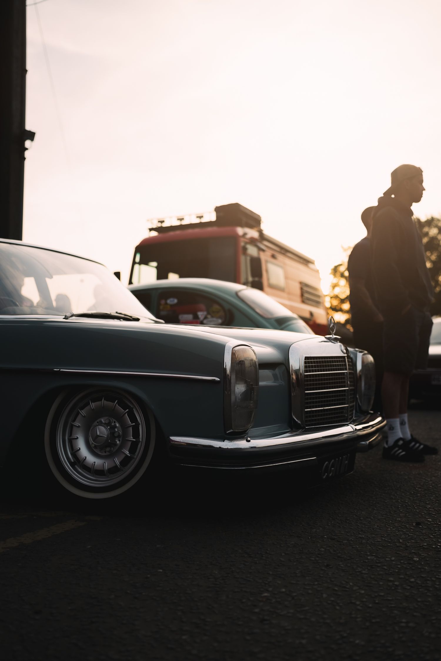 Vintage Mercedes-Benz lowrider car parked at sunset with a silhouetted man standing nearby and a truck in background.