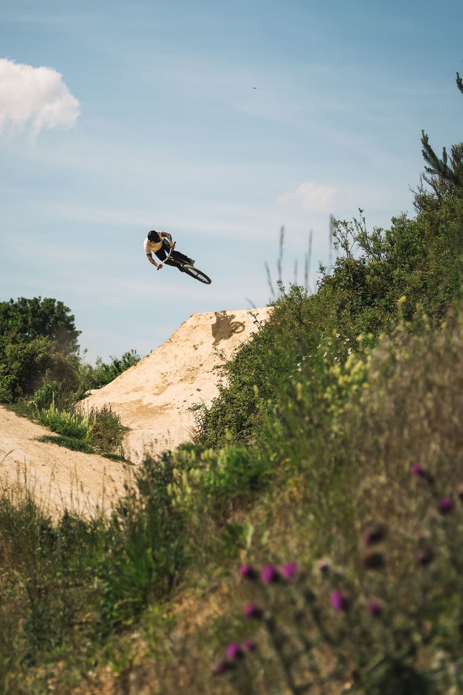 A BMX rider catches big air off a sandy dirt jump surrounded by green vegetation.