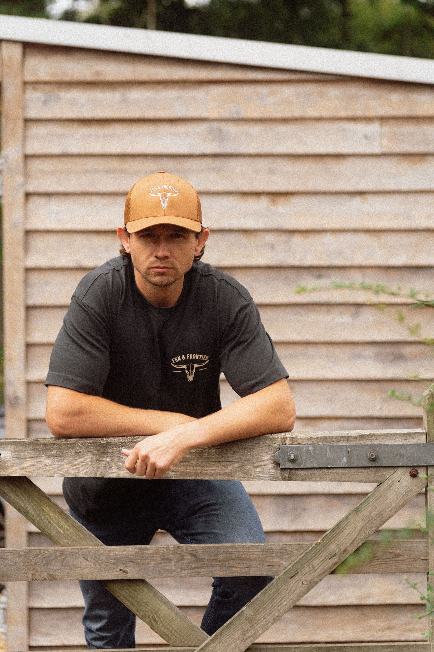Man in brown trucker cap and dark t-shirt leaning on wooden farm gate with rustic shed background.