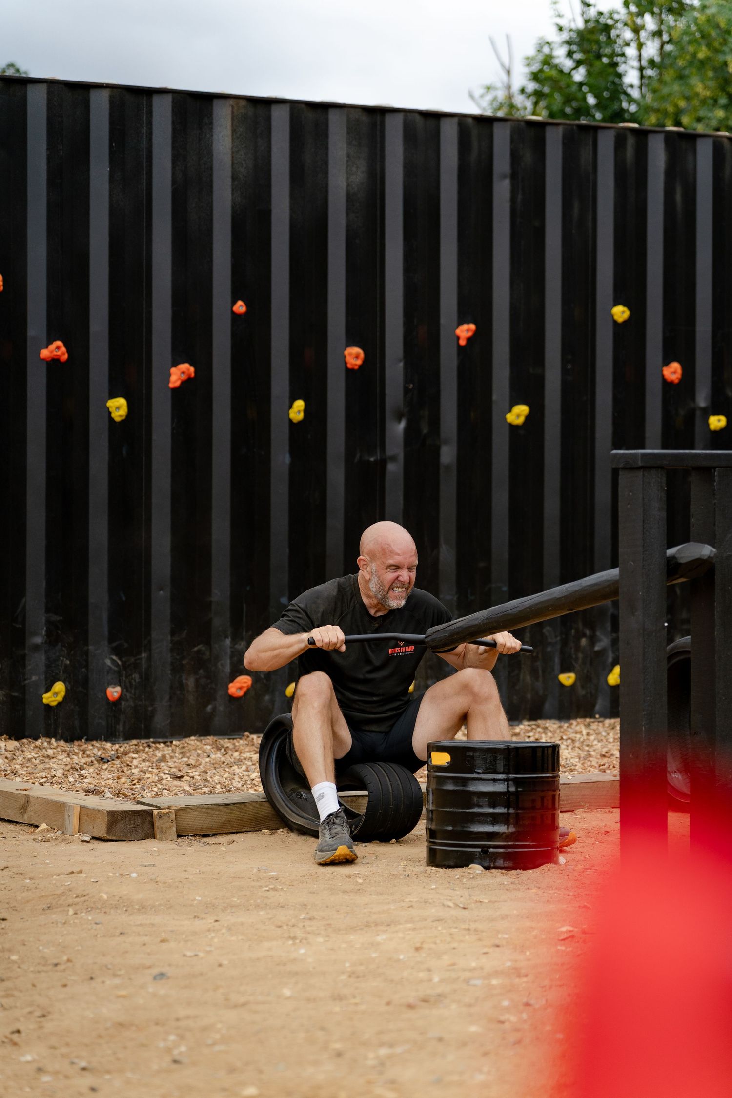 Bald man exercising outdoors at a fitness obstacle course, pulling a weighted sled on sandy ground near a climbing wall.
