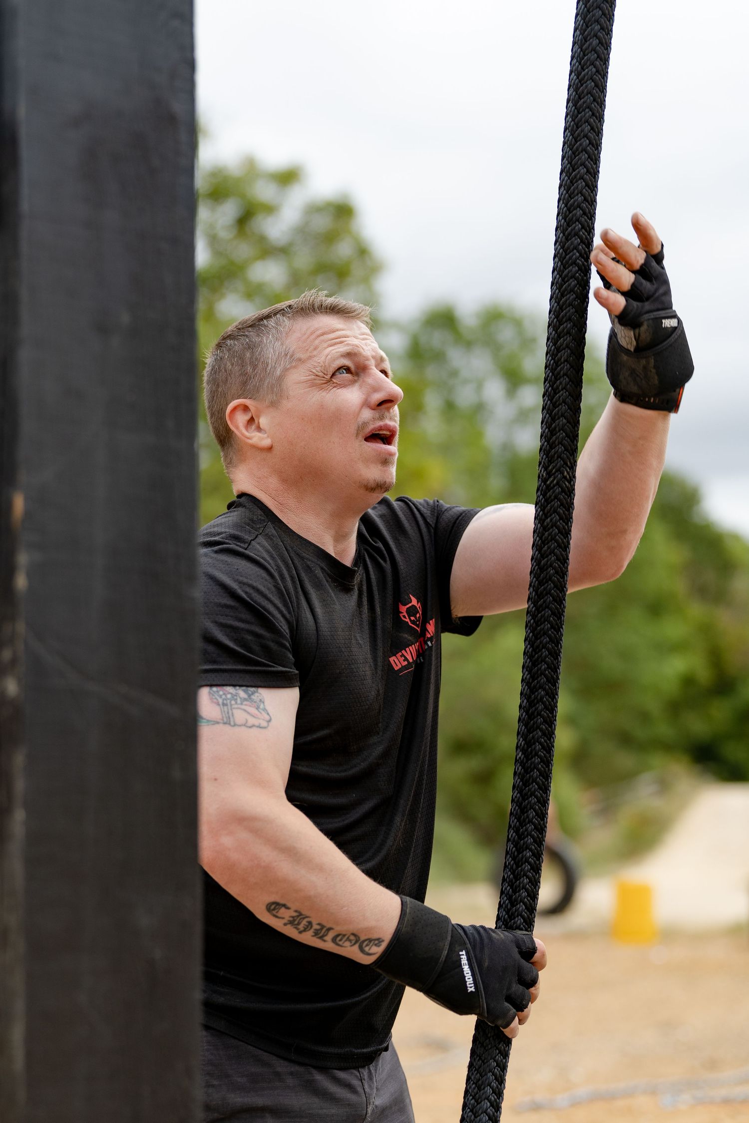 Man in black shirt climbing a rope at an outdoor obstacle course race event, showing determination and strength.