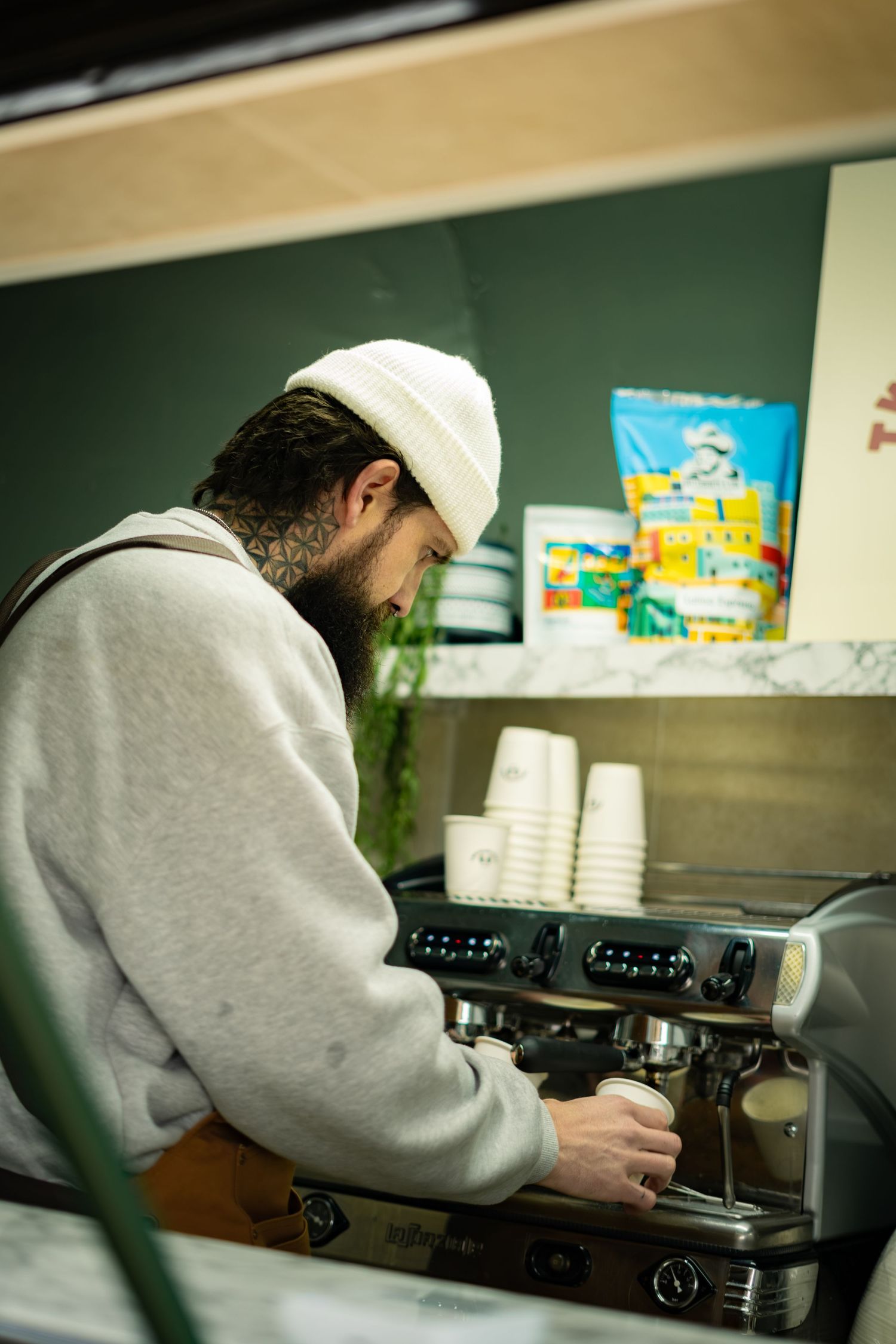 Tattooed barista in white beanie prepares espresso on professional coffee machine in green cafe.