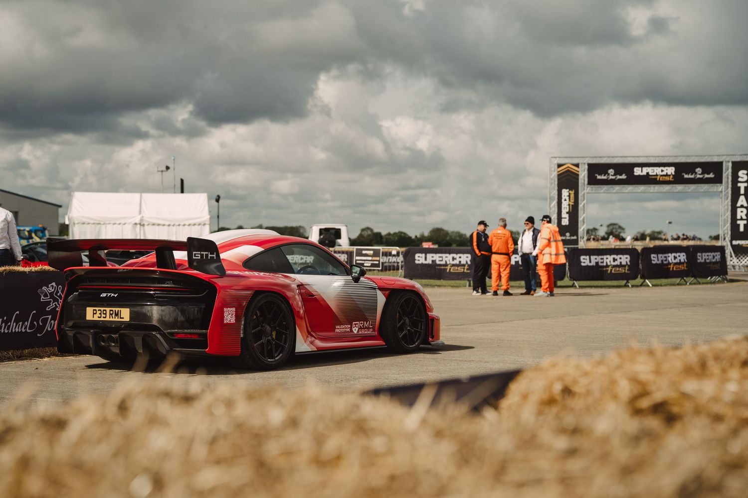 A red McLaren supercar at a motorsport event with hay bales, orange-clad marshals, and cloudy skies in the background.