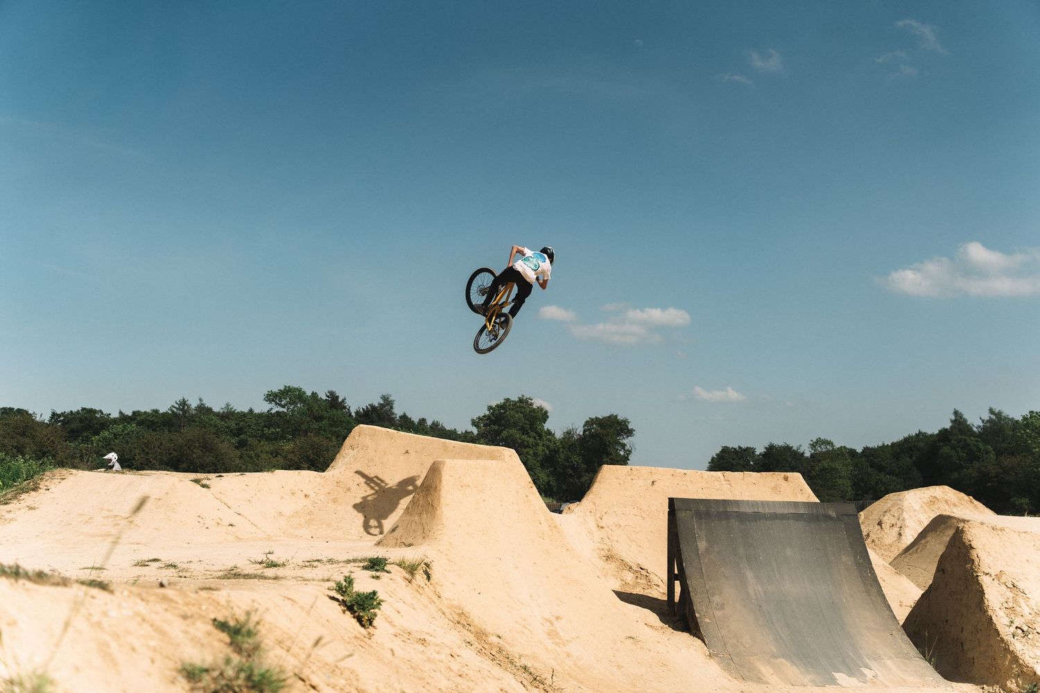 BMX rider performs a high aerial trick off a dirt jump ramp at an outdoor bike park under a clear blue sky.