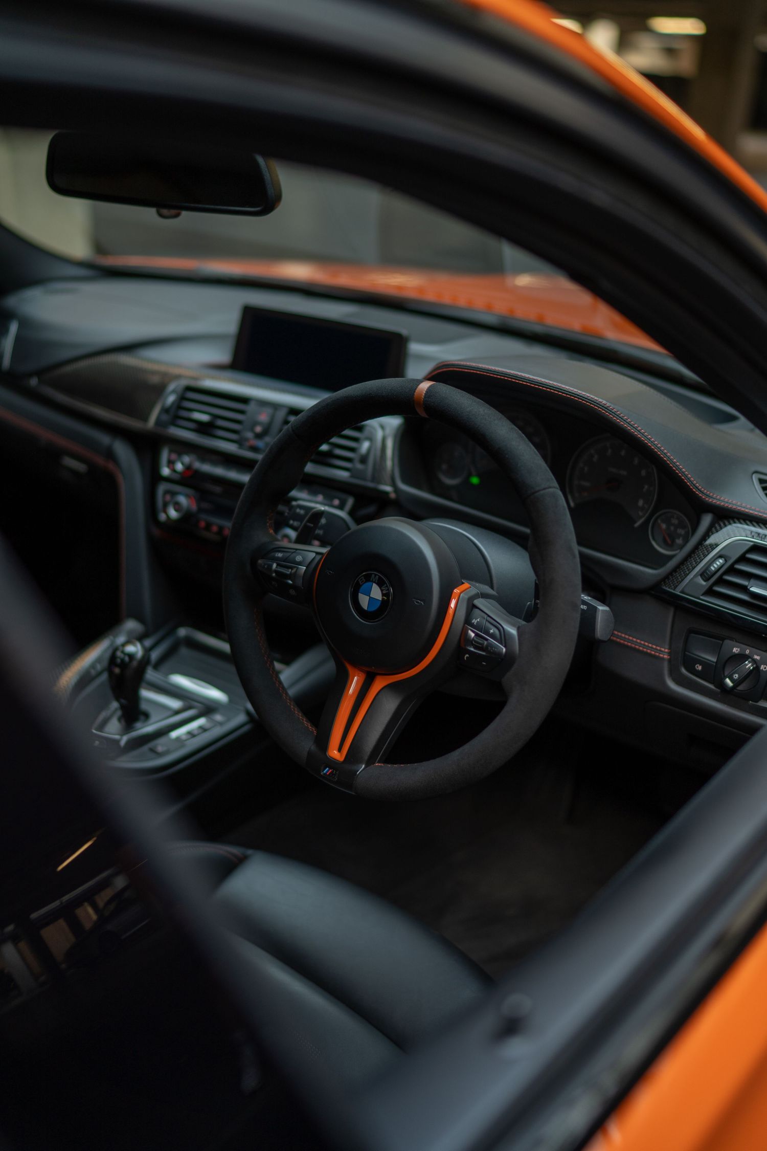 Interior of a BMW M4 with black suede steering wheel featuring orange accents, sport seats, and right-hand drive layout.