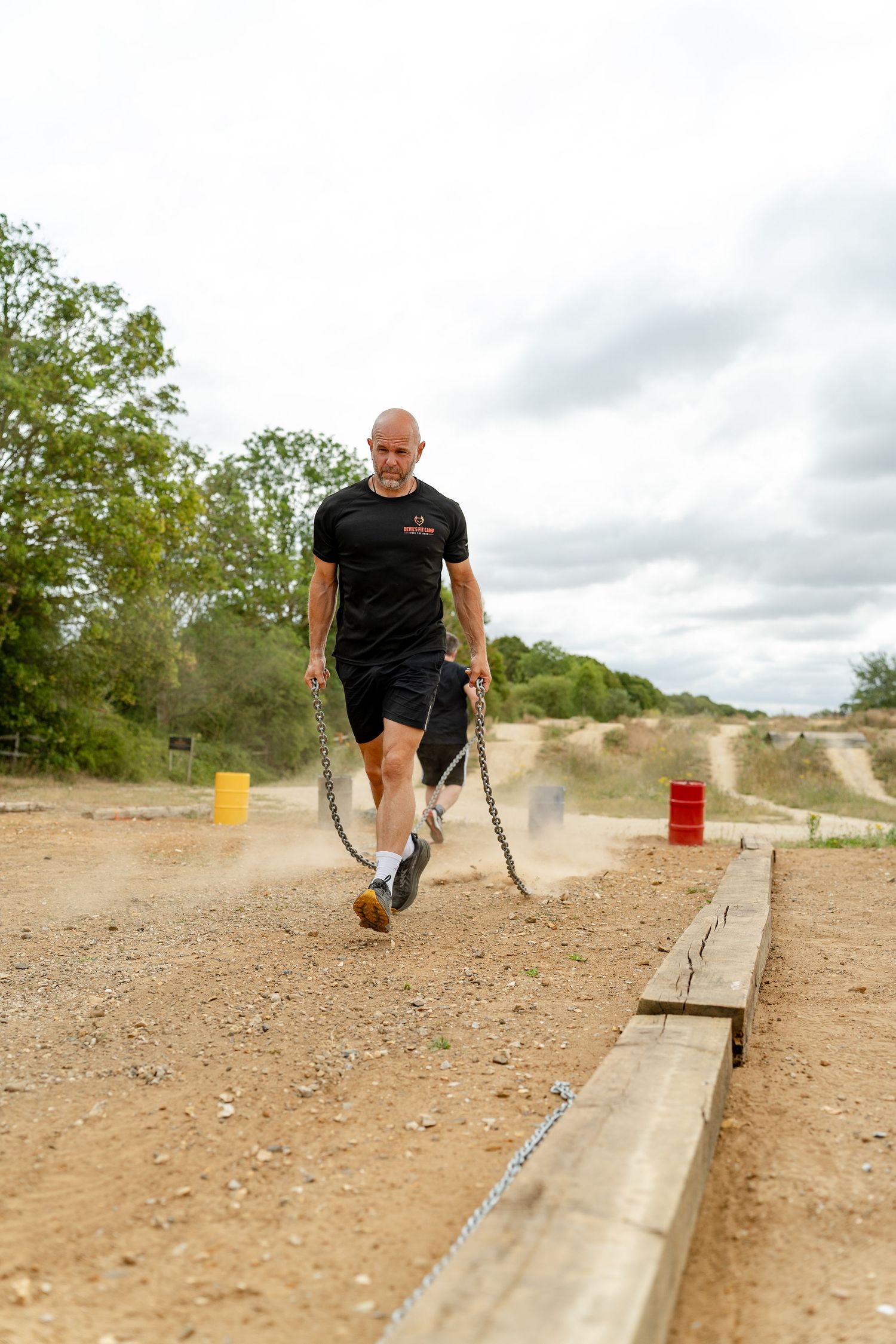 Bald man in black outfit dragging heavy chains across a dirt obstacle course track outdoors.